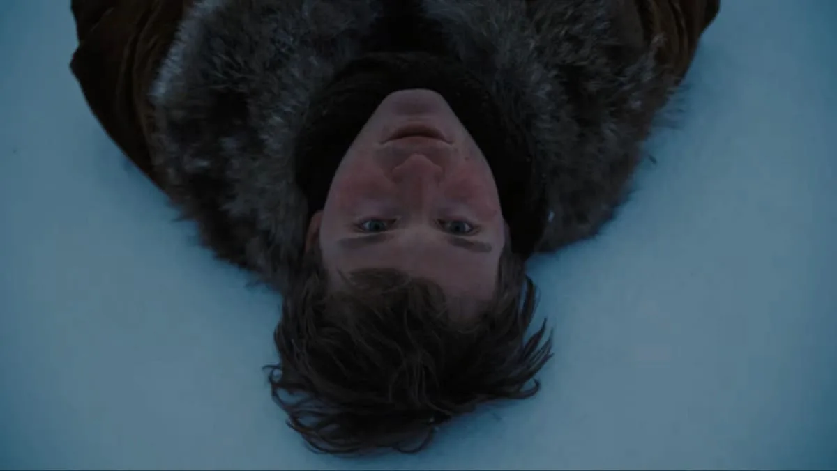 An overhead close-up shot shows actor Fred Hechinger as Mike lying flat on his back against a vast, pristine blanket of blue-tinted snow in the dramatic film A Long Winter. He is wearing a heavy, rustic winter coat with a thick, dark fur collar that frames his face. His head is tilted directly up toward the camera, and his slightly messy dark hair spreads out beneath him on the frozen ground. His expression is one of vulnerable quietude, capturing the raw pain and deep isolation of a young man processing profound grief and navigating an unforgiving winter landscape. An overhead close-up shot captured from a high-angle perspective shows a fair-skinned young person with messy, dark brown hair lying flat on their back on a vast, seamless field of pale blue-white snow, staring directly up into the camera with an intense, wide-eyed gaze. Their head is positioned at the lower center of the frame, inverted from the traditional orientation, with their body extending toward the top. The individual wears a heavy, dark winter cloak with a thick, textured grey and brown fur collar that frames their neck and shoulders, contrasting sharply against the smooth, uniform surface of the snow. The lighting is cold, diffused, and somber, casting soft shadows across the person's face, highlighting their youthful features and the subtle freckles across their nose and cheeks, emphasizing the quiet, atmospheric, and desolate stillness of the frozen landscape.