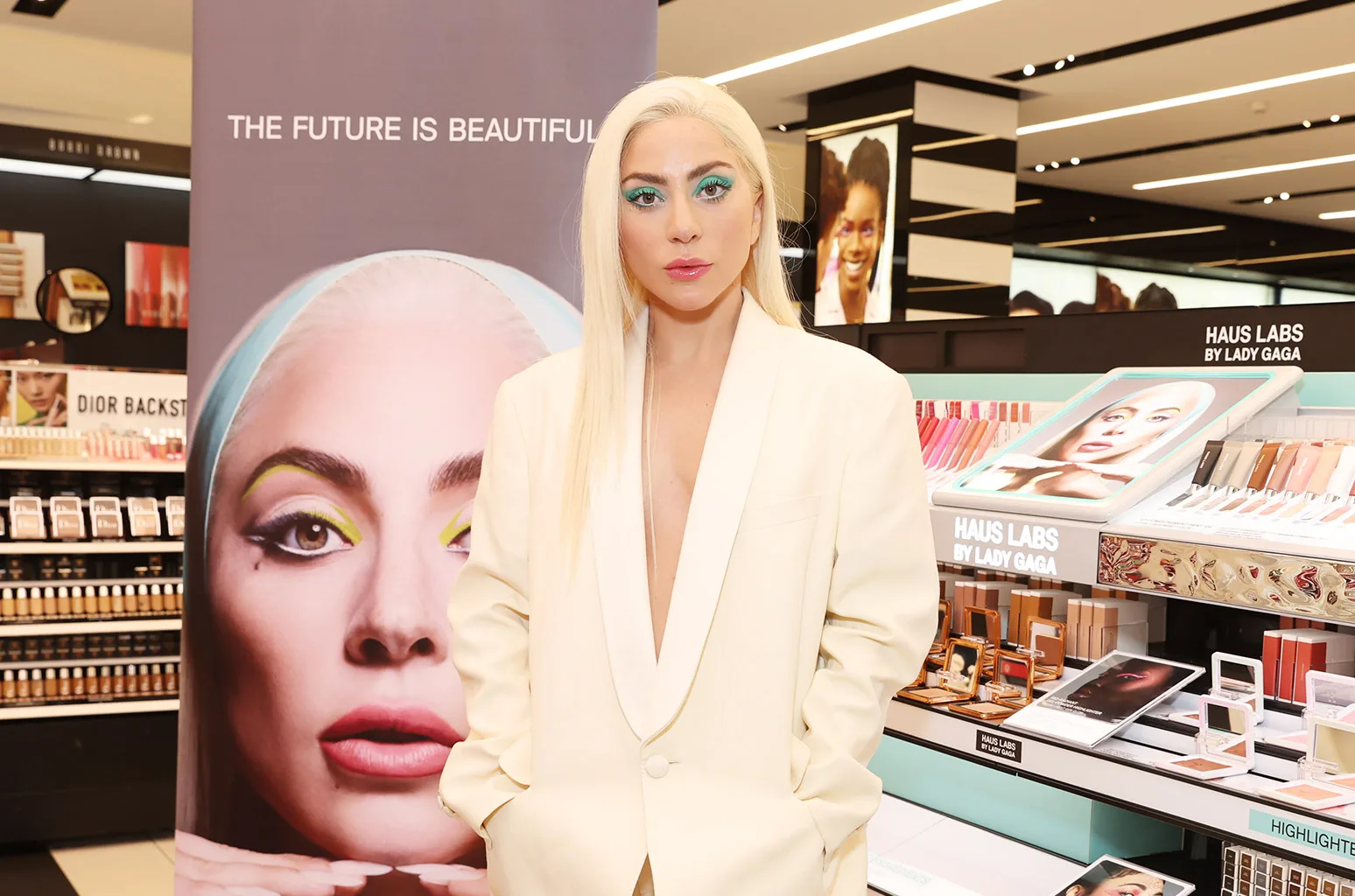 Lady Gaga stands inside a beauty retail store for a brand event, positioned between a large promotional poster of her face and a makeup counter. She is dressed in an oversized, cream-colored blazer with a low-cut satin lapel, worn without a shirt underneath. Her platinum blonde hair is straight, parted down the middle, and falls over her shoulders. Her makeup features a bold teal eyeshadow swept across her eyelids, structured brows, and a soft pink lip. To her right, a tiered beauty display showcases HAUS LABS by Lady Gaga cosmetics, including foundations, lip products, and highlighters. In the background, illuminated shelves, store signage, and a secondary display featuring a model's portrait fill the bright retail environment.
