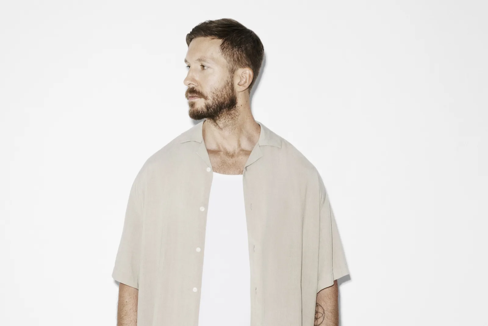 The provided image features a studio portrait of Scottish DJ and music producer Calvin Harris posing against a stark, minimalist white background. He is captured from the waist up, turned in profile facing to his left while sporting short-cropped brown hair and a neatly trimmed beard. Harris is dressed in a relaxed summer outfit consisting of an unbuttoned, short-sleeved beige linen shirt layered over a simple white tank top.