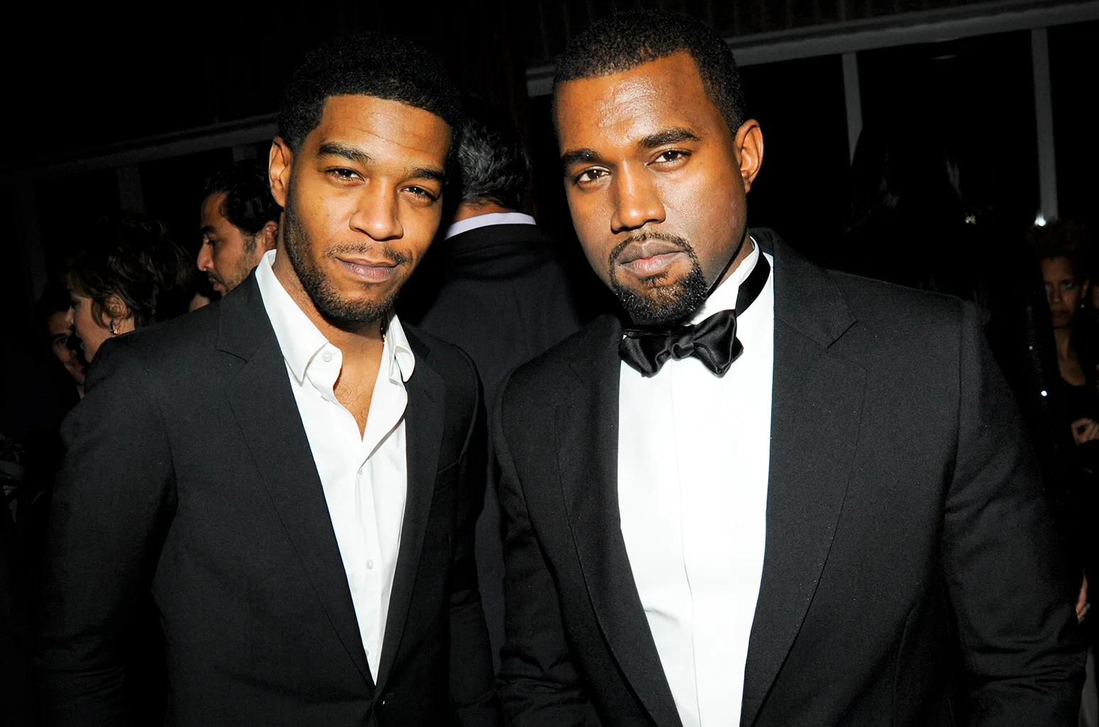 A medium close-up photograph features American musicians Kid Cudi and Kanye West standing side by side against a dark, indoor background at an event. On the left, Kid Cudi smiles subtly at the camera, sporting a trimmed beard and short hair while wearing a tailored black suit jacket over an unbuttoned white dress shirt without a tie. To his right, Kanye West looks directly forward with a serious expression, wearing a sharp black tuxedo jacket, a crisp white tuxedo shirt, and a neatly tied black bow tie. The lighting is direct, casting slight shadows behind them and softly illuminating a few other attendees blurred in the dark background.