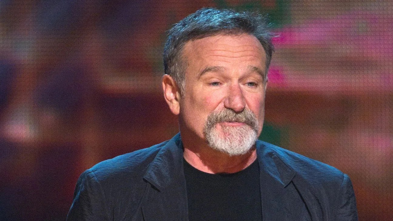 An expressive medium shot captures the actor and comedian Robin Williams from the chest up against a softly blurred, colorful background. He is turned slightly to the right, looking down with a reflective, serious expression. He has short, graying brown hair and a thick, well-groomed gray goatee and mustache. He is wearing a dark, textured charcoal blazer over a simple black crewneck t-shirt. The background features warm, ambient lighting with hints of pink, purple, and green, suggesting a stage or studio setting.