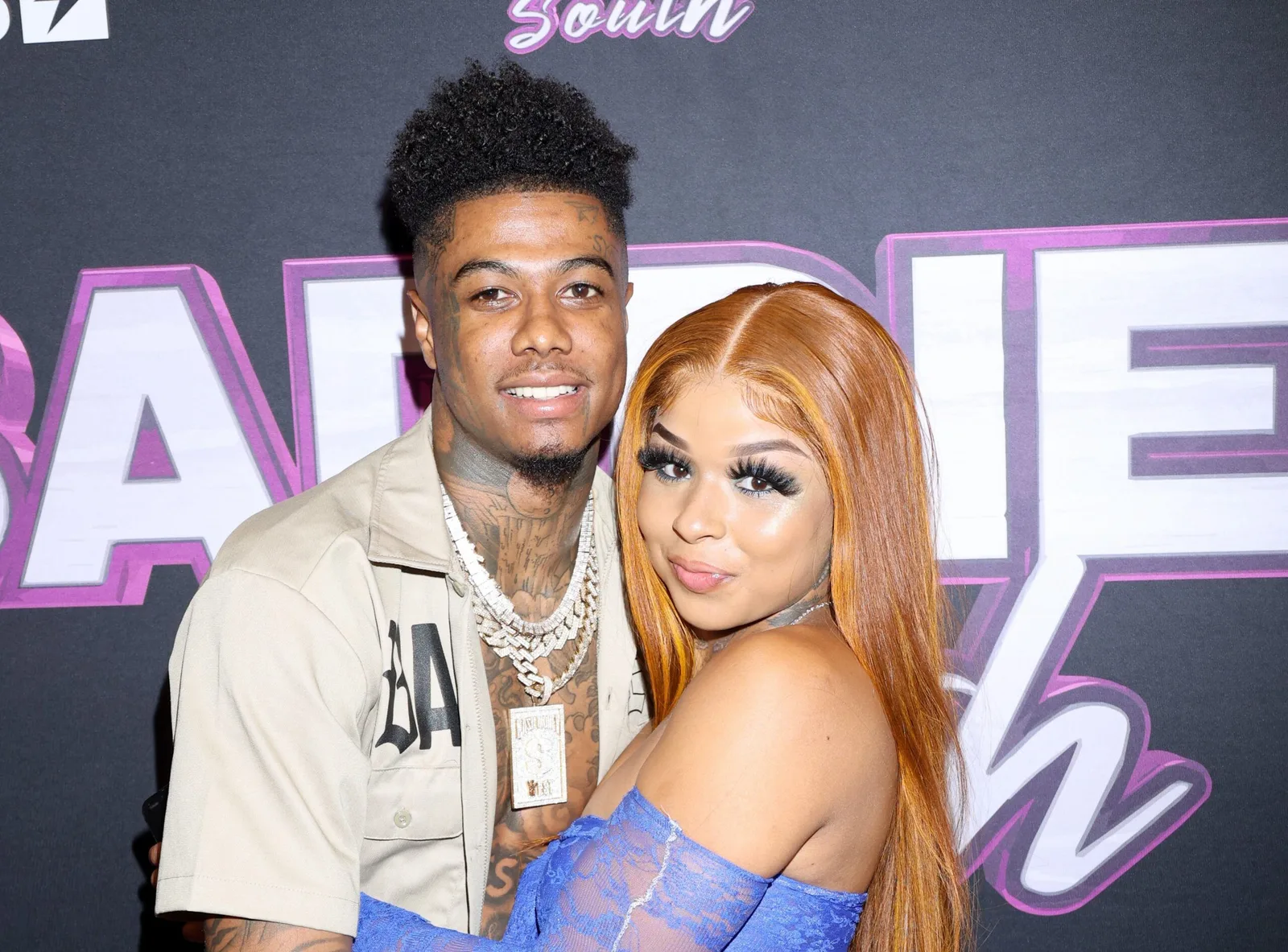 A medium close-up shot captures American rapper Blueface and reality television personality Chrisean Rock posing closely together against a black promotional backdrop featuring large, stylized pink and white lettering. On the left, Blueface smiles forward, sporting a high-top fade haircut, a light beige collared short-sleeve shirt left partially open, and multiple layered thick diamond-encrusted Cuban link chains with a large rectangular pendant. On the right, Chrisean Rock leans against him while looking directly at the camera with a slight smile, showcasing long, straight copper-brown hair, dramatic false eyelashes, and a blue lace off-the-shoulder top.
