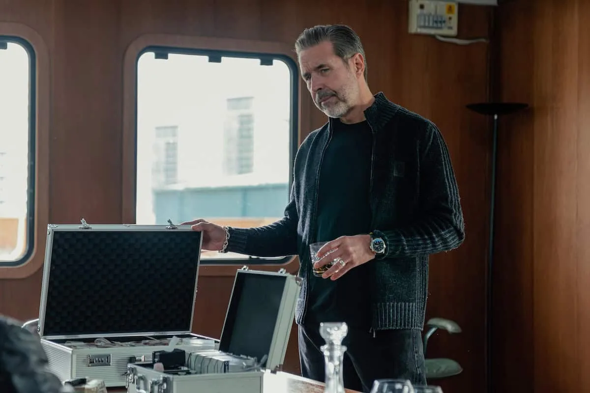 The image shows English actor Paddy Considine playing the character Kevin Harrigan in a promotion for the second season of the crime drama series MobLand on Paramount Plus. Dressed in an all-black casual ensemble featuring a ribbed zip-up mock-neck sweater over a dark crew-neck t-shirt, he is depicted inside what appears to be a ship or yacht cabin with wood-paneled walls and large curved rectangular windows. The character stands behind a table containing opened aluminum utility briefcases filled with bundled money stacks and various analytical tools, casually holding a lowball glass containing an amber spirit like whiskey while peering down with a serious and contemplative expression.
