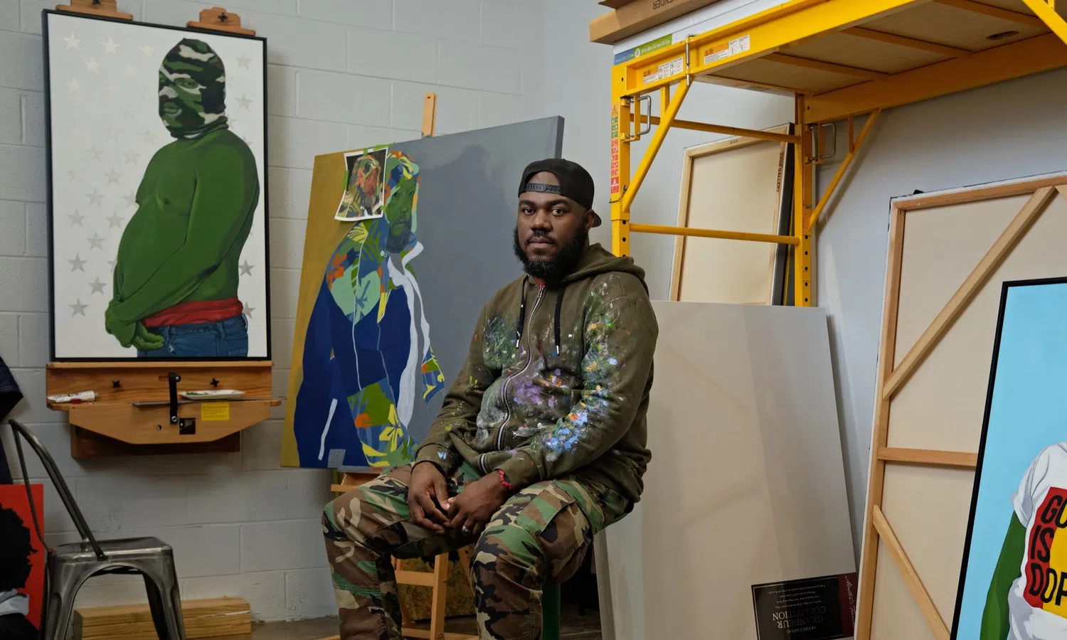 This image features artist Clarence Heyward in his studio surrounded by his works, including the painting God is Dope which depicts a subject with vibrant green skin.