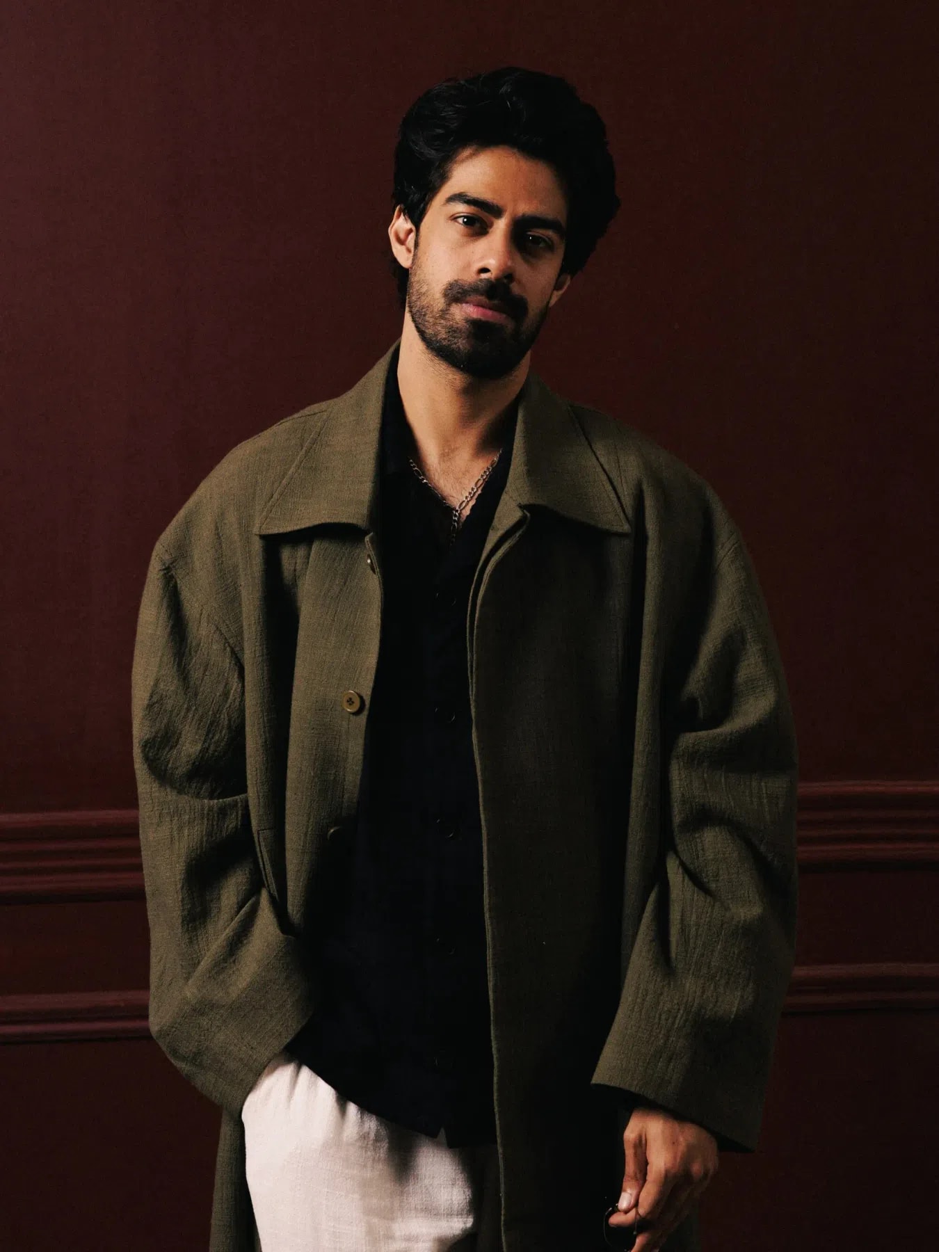 A medium shot of Zain Ahmad looking directly at the camera against a dark red paneled wall. He has thick black hair, a groomed beard, and a mustache. He is wearing an oversized olive green linen trench coat layered over a black button-up shirt that is slightly open at the collar, revealing a silver chain necklace. His hands are loosely placed near the pockets of his off-white or light beige linen trousers, creating a relaxed yet fashionable posture.
