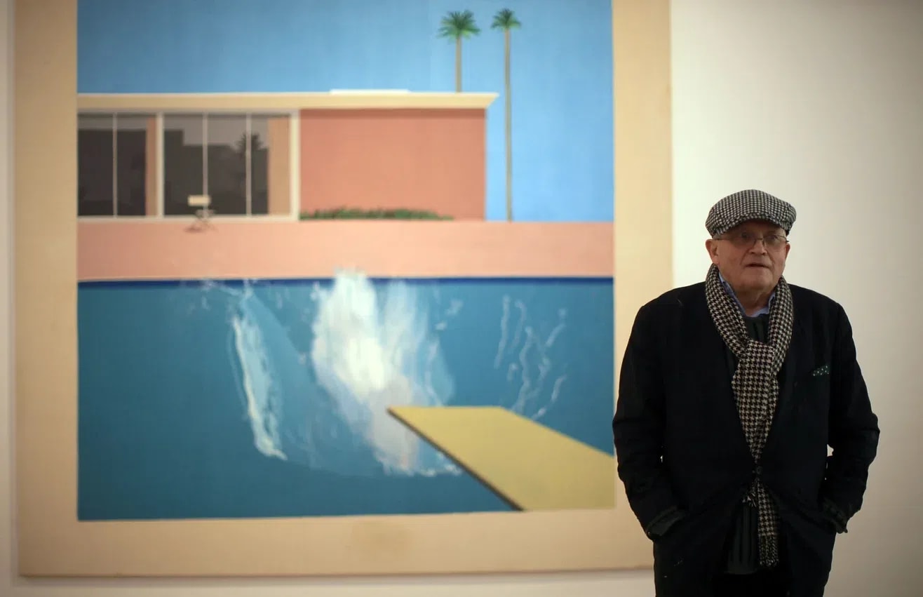 A wide, eye-level shot captures British artist David Hockney standing to the right of his famous large-scale pop art painting, A Bigger Splash (1967), which is mounted on a plain white gallery wall. The painting itself takes up the majority of the left and center portions of the frame. It depicts a modernist, flat-roofed house with floor-to-ceiling glass windows under a bright, clear blue sky, flanked by two tall, thin palm trees. In the foreground, a yellow diving board extends from the bottom right into a swimming pool of turquoise water, where a massive, chaotic splash of white and light blue water erupts, indicating someone has just dived in. The entire painting is bordered by a wide, unpainted canvas margin. To the right, David Hockney stands in a three-quarter view, looking slightly past the camera to the left. He is an elderly man with glasses, wearing a dark navy blazer, a black and white houndstooth scarf draped around his neck, and a matching houndstooth flat cap. His hands are tucked into his pockets, and his expression is neutral and contemplative. The lighting is soft and even, typical of a gallery setting, casting subtle shadows behind him.