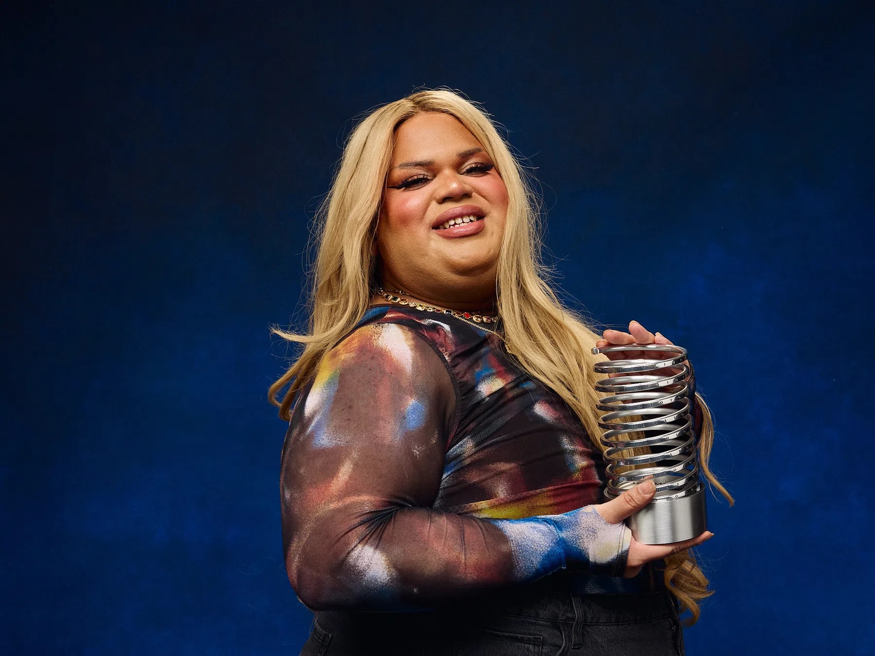 Jools Lebron is an American TikTok creator and beauty influencer who achieved massive, overnight internet fame in August 2024 after coining the viral satirical phrase "very demure, very mindful". A transgender Latina woman of Puerto Rican heritage from Chicago, Lebron was working as a supermarket cashier and creating makeup tutorials before her video humorously detailing how to present oneself modestly in the workplace exploded into a global pop-culture phenomenon. The catchphrase took over social media, was adopted by countless major brands and celebrities, earned her a guest spot on Jimmy Kimmel Live!, and was ultimately named Dictionary.com's Word of the Year. Despite her rapid success—which initially allowed her to fund her gender transition and secure brand partnerships—the sudden lifestyle shift and overwhelming pressure of viral fame severely impacted her mental health, leading to public struggles with impulsive spending and substance abuse. Recently, Lebron faced significant internet backlash after launching a $45,000 GoFundMe campaign to repair dental rot caused by her past drug addiction. Though she quickly canceled the fundraiser and refunded her donors due to the criticism, the intense public scrutiny led her to announce a step back from TikTok to focus on her sobriety, recovery, and mental well-being away from the spotlight. A half-length studio portrait of social media creator Jools Lebron smiling warmly against a dark blue background. Lebron, a trans woman with long blonde hair, bright makeup, and bold eyeliner, is wearing a long-sleeved mesh top featuring a dark, colorful abstract print. She is standing in profile, turned toward the right side of the frame while looking back at the camera. In her hands, she proudly holds a silver, metallic trophy shaped like a large coil spring with a solid metal base. 