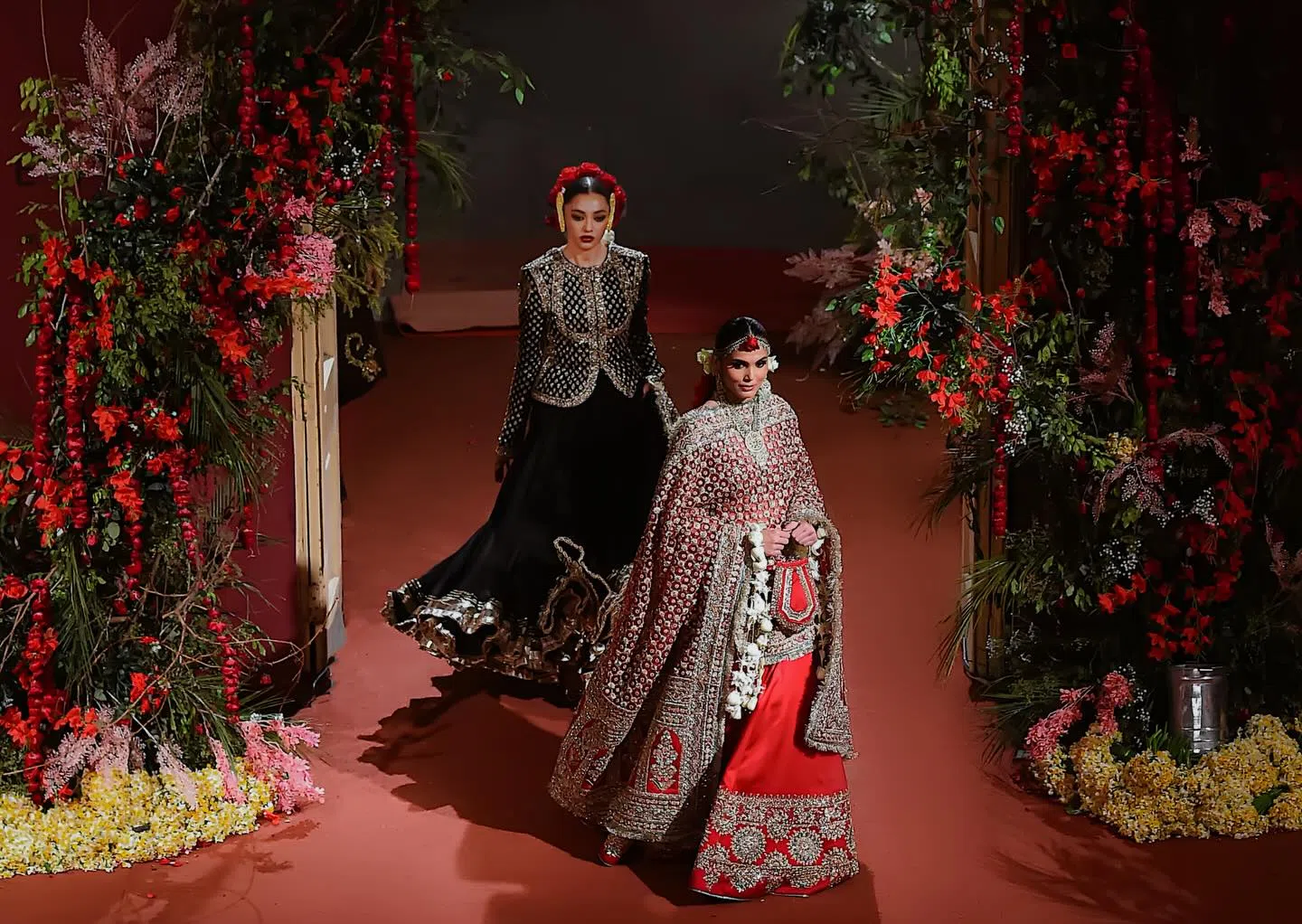 A wide, high-angle shot captures two models presenting the "Nargis" Bridal Couture SS 26 collection on an indoor runway floored with solid terracotta-red carpeting.Foreground Model: Walking confidently forward on the right, she wears a heavily traditional red bridal lehenga. The ensemble features a flowing crimson skirt hemmed with thick gold embroidery and a matching, heavily draped shawl-style dupatta covering her torso and arms. Her styling includes a traditional maang tikka, dangling earrings, a sleek middle-parted low bun, and an elaborate, dangling white-flower kiran garland clutched alongside a small bag.Background Model: Walking slightly behind and to the left, she models an elegant jet-black lehenga choli. The outfit comprises a structural, full-sleeved black jacket-style choli covered in geometric gold zari grids and a wide, layered black skirt that flares dramatically toward the hem with matching gold trimmings. A crown of bright red roses accents her slicked-back hairstyle.Framing the runway are massive, asymmetrical floral installations. Dense arrangements of lush green foliage, trailing vines, and blooming crimson and soft-pink roses grow along the sides. Scattered yellow and white flower petals blanket the base of the arrangements.