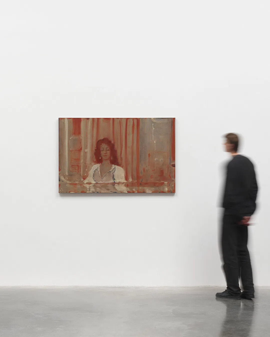 An installation photograph from White Cube Paris showcases a rectangular, horizontally oriented figurative painting by Pakistani artist Suleman Aqeel Khilji. The artwork is mounted centrally on a large, pristine white gallery wall. Executed with a muted, warm palette dominated by earth-toned reds, terracottas, and hazy greys, the dreamlike composition depicts a woman with voluminous dark curly hair sitting behind what appears to be a counter or reflective surface. She wears a white, low-cut blouse or dress with dark trim along the neckline. The background features hazy vertical bands resembling curtains or architectural pillars, rendered in semi-translucent layers of pigment that evoke a sense of a faded memory or an excavated artifact. To the right of the canvas, a person wearing a black long-sleeved shirt and dark trousers stands on a polished concrete gallery floor, captured in motion blur as they look toward the left.