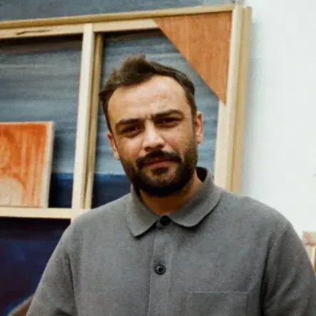 An average-looking adult man (Suleman Aqeel Khilji) with tanned skin, a neat dark beard, and short hair stands in a brightly lit art studio, looking directly at the camera with a gentle, relaxed expression. He is wearing a grey buttoned long-sleeve shirt jacket with a pocket on the left breast and dark grey pants. He is standing in the middle of the frame, surrounded by large, unframed paintings. Behind him on the left, a large canvas stretcher leans against the wall, revealing smaller rectangular canvas paintings peeking out. The surrounding artwork features earthy tones of brown, tan, and blue, depicting abstract landscapes and figures. The studio has a light-colored floor and white walls.