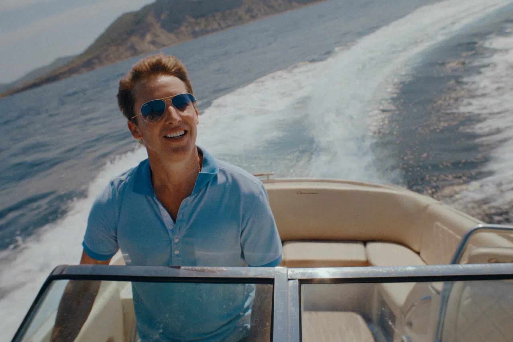 English singer-songwriter James Blunt smiles brightly while piloting a motorboat through deep blue open waters, leaving a massive white sea spray wake trailing behind him. He wears a light blue polo shirt with darker blue trim on the collar and sleeves, paired with classic metallic aviator sunglasses that reflect the coastal surroundings. Behind him, a rugged, mountainous coastline rises out of the ocean under a clear, bright sky, framing the sun-drenched, high-speed maritime scene.