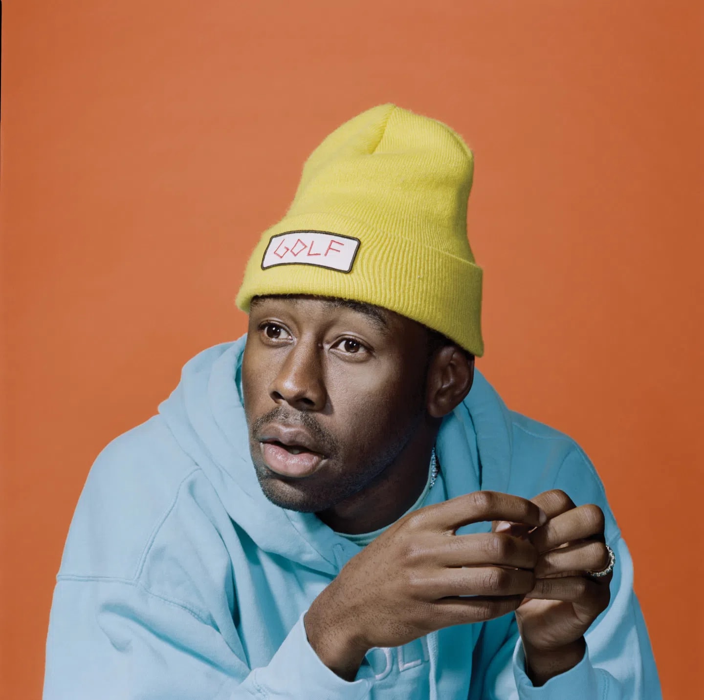 A close-up portrait of the artist Tyler, The Creator against a solid, vibrant orange backdrop. He is wearing a bright yellow knit beanie adorned with a white rectangular patch featuring the word "GOLF" in pink, stylized lettering. He sports a light blue hoodie with embroidered text partially visible near the collar. His expression is contemplative with an open-mouthed, slightly surprised look as he gazes off-camera to his left, his hands raised in front of him with a silver ring visible on his pinky finger.