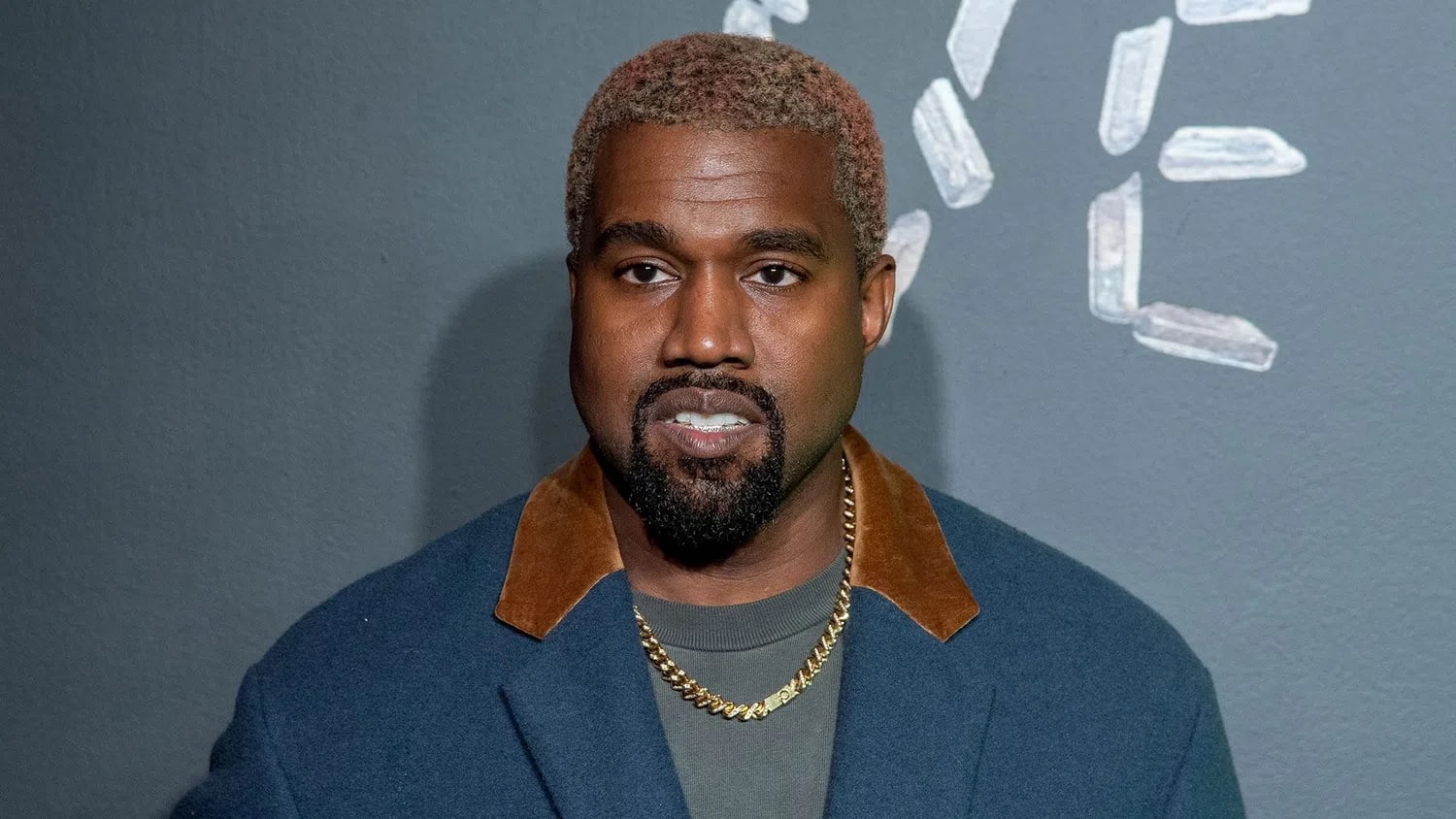 A close-up portrait shows American rapper, fashion designer, and entrepreneur Kanye West (officially known as Ye) looking directly into the camera while standing against a dark grey textured wall accented with scattered clear crystal formations. He is styled with short, buzz-cut hair dyed in faded pink and blonde tones, a neatly trimmed goatee, and a thick gold chain necklace around his neck. He wears a dark grey crewneck undershirt layered beneath a dark blue wool blazer that features distinctive brown suede or velvet collars.