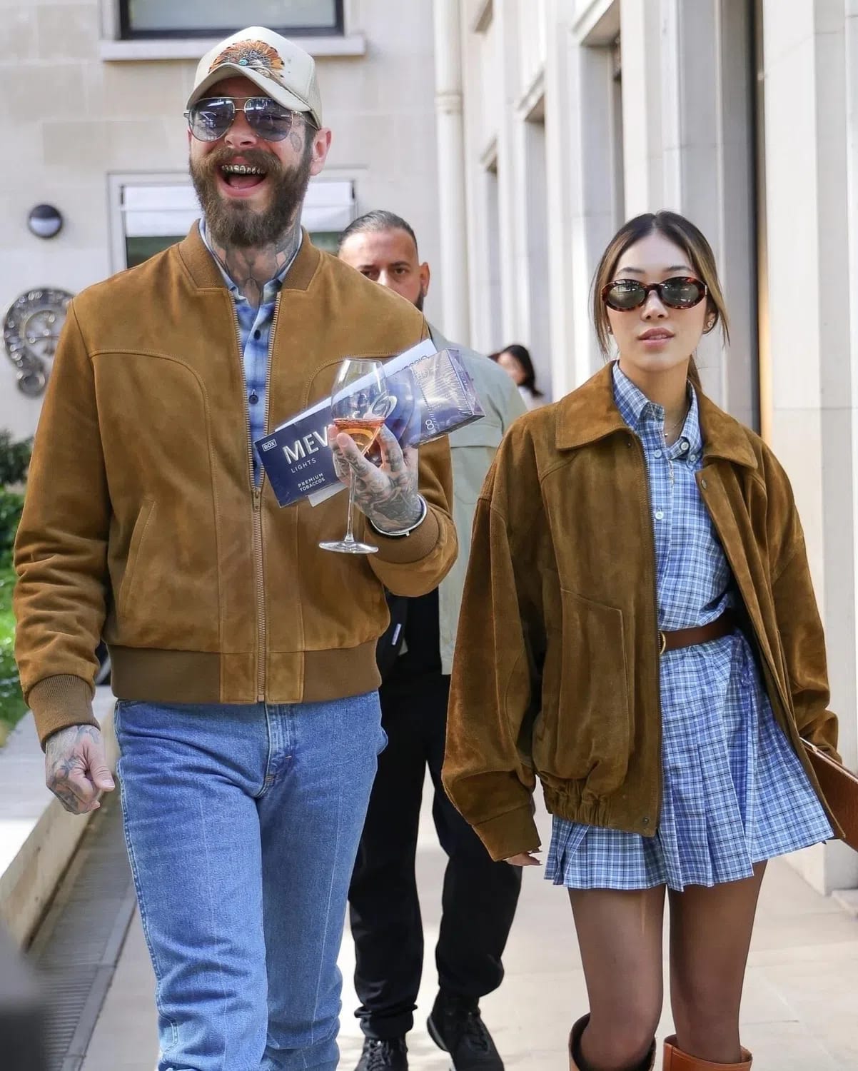 An image shows American musician Post Malone laughing while walking outdoors alongside his partner, Christy Lee, both wearing matching brown suede jackets over blue patterned tops. Post Malone, on the left, is wearing a structured brown suede bomber jacket over a blue and white checkered shirt, paired with light-wash blue jeans, a light-colored baseball cap, and aviator sunglasses, while holding a wine glass and a blue box of Mevius Lights cigarettes. Christy Lee, on the right, wears a loose-fitting brown suede jacket over a blue plaid mini dress secured with a brown leather belt, styled with tortoiseshell sunglasses and tall brown leather boots, as they walk down a paved pathway flanked by light-colored building facades with a security guard following closely behind. Post Malone and Christy Lee walk together in coordinating suede outfits. Photo by Backgrid.