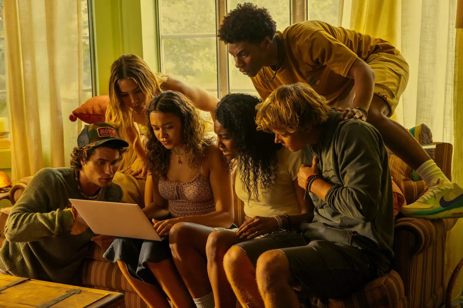 A group of six teenagers from the Netflix series Outer Banks are gathered tightly together in a warm, yellow-toned living room, looking intensely at a silver laptop screen. On the far left, a young man wearing a trucker hat sits on the floor leaning inward. Next to him, a young woman with wavy brown hair holds the laptop on her lap. Behind them stands another young woman leaning over the back of the sofa. In the center, a young woman with dark curly hair watches the screen closely, while a young man with shaggy blonde hair sits next to her, leaning his torso forward. Above them, a young man in a yellow shirt and shorts climbs over the back of the sofa, leaning completely over the group to catch a glimpse of the screen.