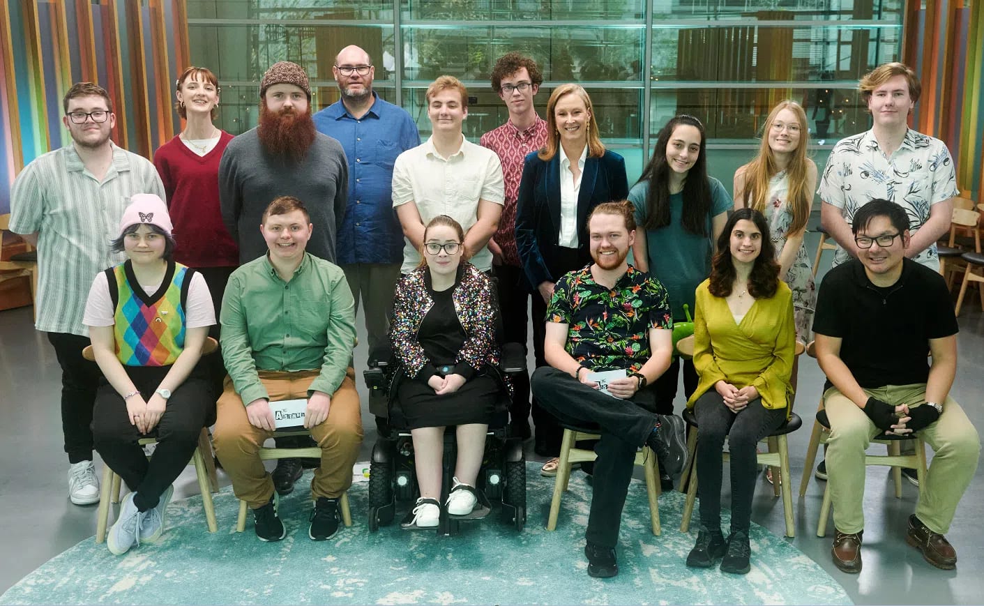 A group portrait of the cast and crew from the Australian documentary series, The Assembly. The group consists of fifteen individuals arranged in two rows inside a modern room with a backdrop of colorful, vertical stripes. In the back row, eight people stand side by side: a man in a light green striped shirt, a woman in a red V-neck sweater, a man with a large red beard wearing a grey sweater, a tall man with glasses in a blue button-down shirt, a man in a white polo shirt, a man in a patterned pink shirt, host Leigh Sales in a dark blue velvet blazer over a white shirt, a woman with long dark hair, a woman with long blonde hair, and a man in a white short-sleeve shirt with a leaf pattern. In the front row, six individuals sit facing forward: a person with colorful rainbow argyle vest and pink beanie, a man in a green shirt and khaki pants holding a sign, a woman with glasses and a sparkling jacket sitting in a motorized wheelchair, a man in a tropical floral shirt, a woman in a yellow wrap top, and a man in a black polo shirt and beige pants wearing arm compression sleeves.