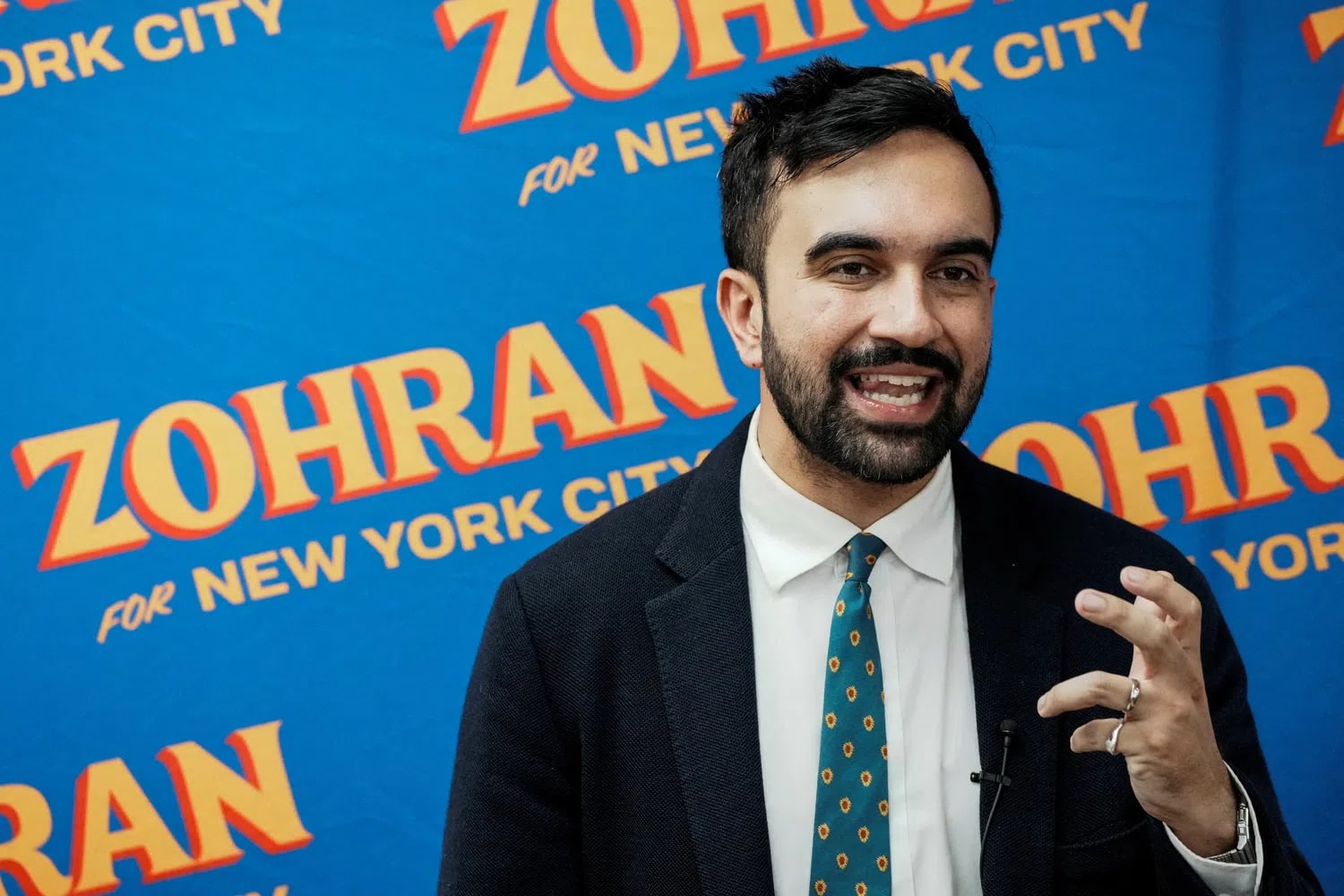 A man with short dark hair and a beard stands in front of a blue background that features a repetitive orange and yellow logo reading "ZOHRAN FOR NEW YORK CITY." He is wearing a dark blazer over a white collared shirt paired with a green tie that has a small, repeating patterned design. He is captured mid-speech, looking to his right with his mouth open and his left hand raised, fingers partially curled, as if gesturing to emphasize a point.