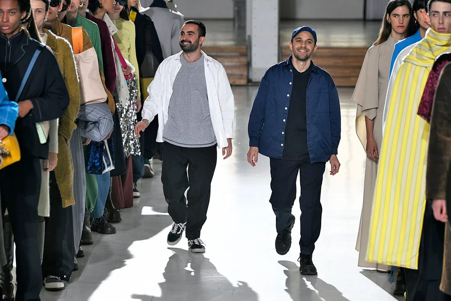 A medium full shot captures fashion designers Loris Messina and Simone Rizzo walking forward down a clean white runway during a fashion show. Messina, on the left, is smiling and has a dark beard and short hair. He wears an open white button-up shirt over a heather grey t-shirt, loose-fitting black trousers, and black-and-white low-top Vans sneakers. Rizzo, on the right, smiles while wearing a blue baseball cap, an open navy blue button-up worker jacket over a plain black t-shirt, dark blue trousers, and dark chunky shoes. On both sides of the runway, models are lined up facing inwards, showcasing various colorful designs including a bright yellow and white striped cloak on the right, and metallic sequined fabrics and structured coats on the left. The scene is brightly lit with a smooth, reflective floor.