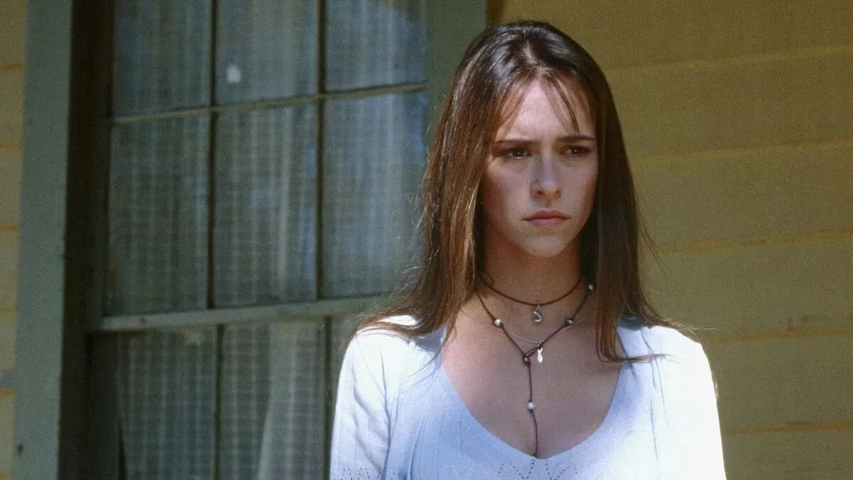 A medium shot captures a young woman with a concerned, somber expression, played by Jennifer Love Hewitt as Julie James in the 1997 horror film I Know What You Did Last Summer. She stands outdoors against a light-yellow wooden wall, with a large, multi-paned window featuring white lace curtains visible on the left. She has long, straight brown hair with wispy bangs framing her face, and is wearing a light blue or white knit top with a wide neckline. Layered around her neck are two thin necklaces: a shorter black cord choker with a small silver charm, and a longer delicate necklace adorned with small pearls and a hanging silver fish pendant that drops down her chest. The lighting is soft and natural, emphasizing her dark eyes and furrowed brow.