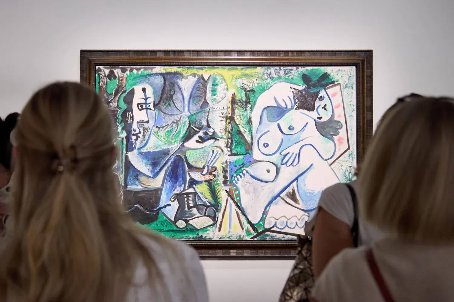 A medium shot captures museum visitors from behind as they observe a large modern art painting hanging on a white gallery wall. The oil-on-canvas artwork, rendered in a late Cubist style, features vibrant shades of green, blue, yellow, and white. On the left side of the canvas, a stylized painter is depicted seated at an easel with brushes in hand. On the right side, a distorted, reclining female nude serves as the model. The painting is framed in a dark, structured wooden border. In the foreground, the blurred back of a woman's head with blonde hair tied in a half-up ponytail fills the lower-left section, while another visitor's blonde bob-cut head anchors the lower-right side.