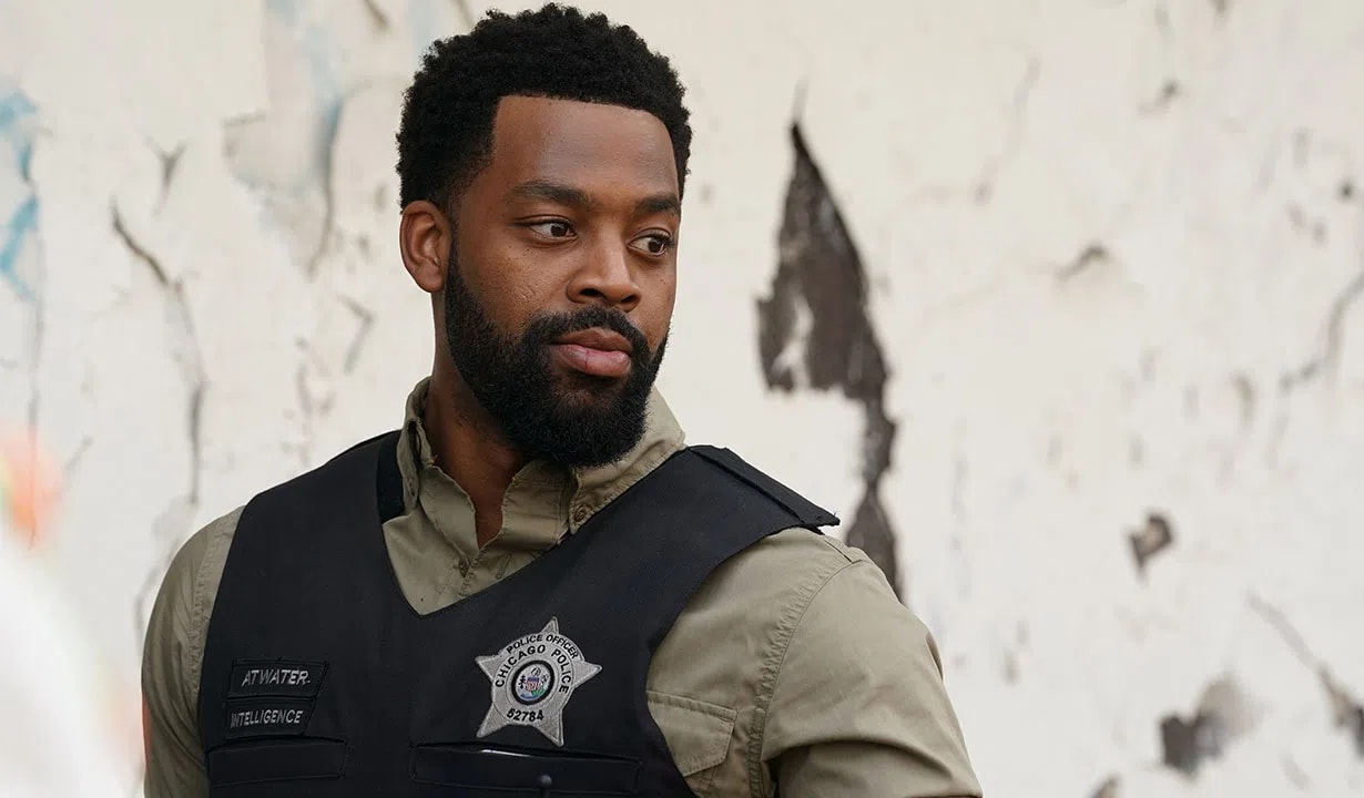 A medium shot of Officer Kevin Atwater, played by LaRoyce Hawkins, from the television series "Chicago P.D." He is standing outdoors against a weathered, peeling white wall, looking off to the side with a serious and focused expression. He wears an olive green button-down shirt under a black tactical police vest. The vest features a silver Chicago Police star badge with the number 52784 pinned to the chest, alongside nametags that read "ATWATER" and "INTELLIGENCE."