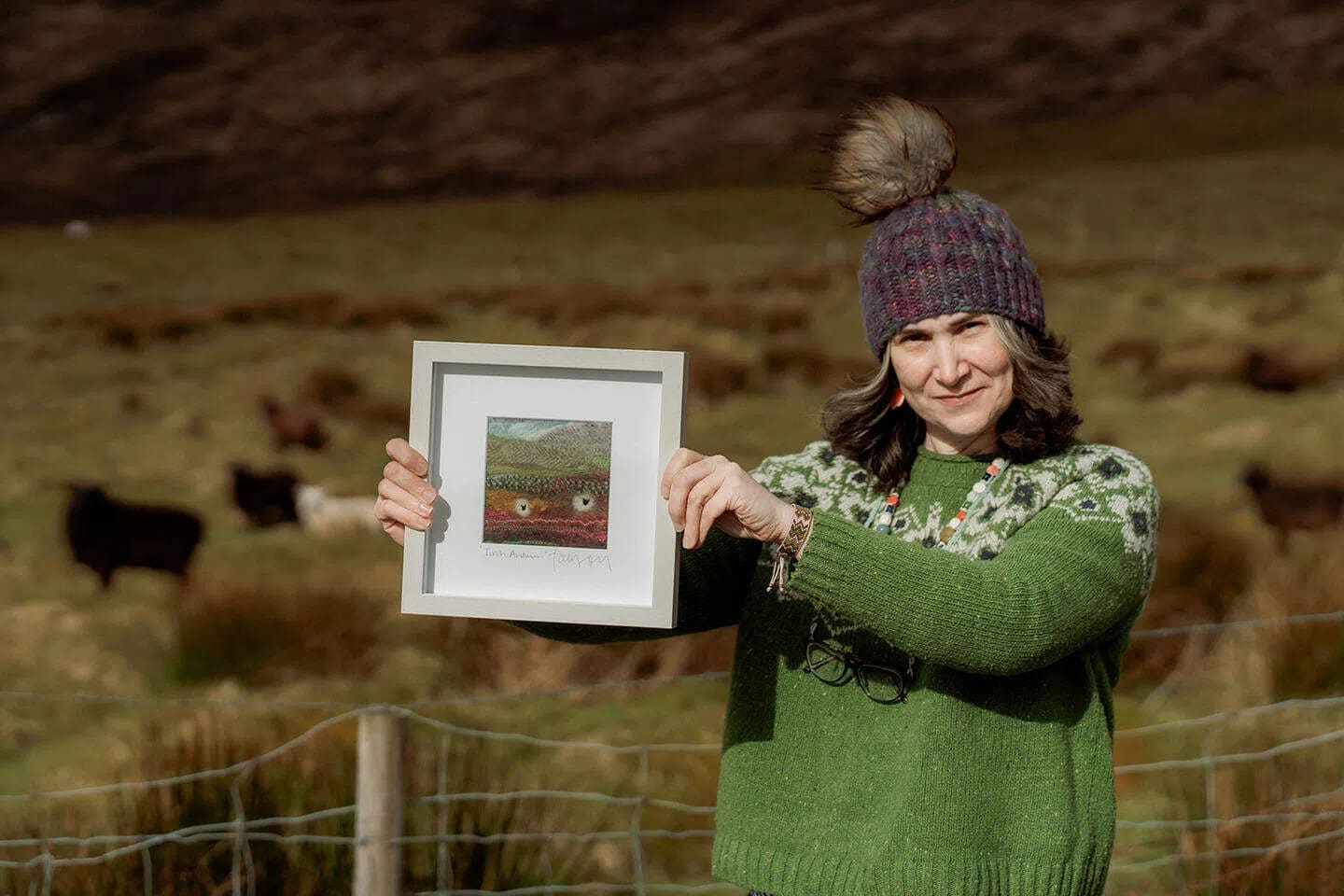 A medium-shot photograph captures a fair-skinned woman standing outdoors in a rural, hilly landscape, smiling gently as she holds up a small, square, light-gray framed artwork. She is positioned on the right side of the frame, wearing a thick, vibrant green knit sweater with a white and dark green Fair Isle patterned yoke, a multi-colored knit beanie with a large faux-fur pom-pom, and dark-rimmed eyeglasses tucked into the front of her collar. Her shoulder-length brown and gray hair frames her face, and she wears long, colorful beaded necklaces. With both hands, she displays the framed piece of art, which depicts a colorful, abstract textile landscape featuring small sheep figures against a hillside, complete with a signature at the bottom. The background consists of a sloping, grassy brown and green hill under bright, natural daylight, with several dark sheep grazing in the distance behind a wire mesh fence with wooden posts that runs along the bottom foreground.