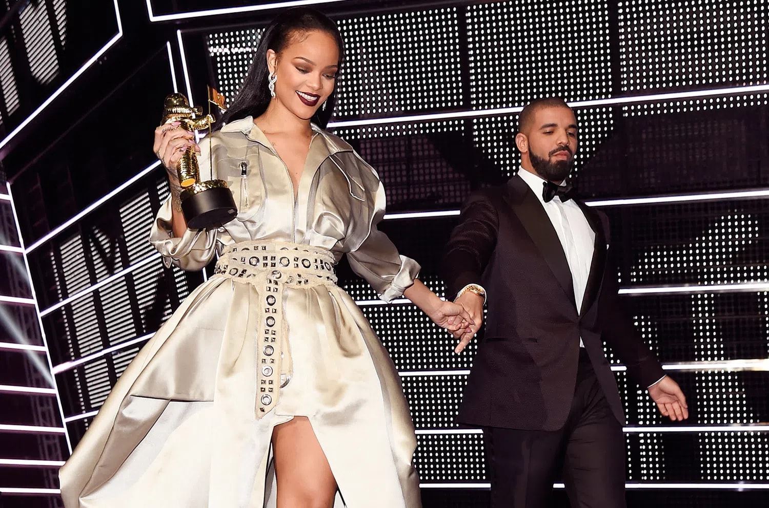 A medium-shot photograph showcases Rihanna and Drake on stage under bright concert lighting. On the left, Rihanna wears an elegant, collared, metallic silver-champagne gown structured like a trench coat, featuring a dramatically slit, flowing skirt and an intricately patterned, wide grommeted belt cinched at her waist. Her hair is styled sleek and straight behind her shoulders, and she wears dramatic dark maroon lipstick while looking downward with a smile. In her right hand, she holds a golden MTV "Moonman" trophy aloft, and with her left hand, she holds Drake’s hand. Drake stands to the right, dressed in a sharp black tuxedo with a crisp white shirt and a black bow tie, gazing toward her with a slight smile. The background features a large grid of geometric, black-and-white LED lighting, adding a modern, high-contrast texture to the scene.