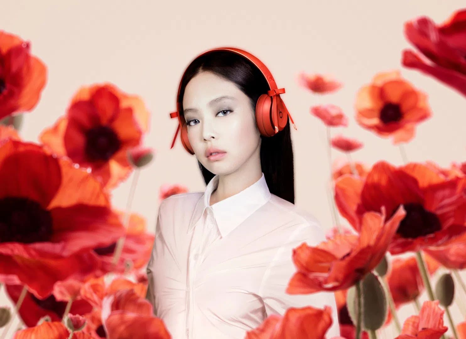 A professional promotional photograph featuring K-pop artist Jennie Kim positioned in the center, looking directly at the camera with a neutral expression. She has straight, dark hair, subtle makeup, and is wearing bright red Beats Solo 4 on-ear wireless headphones that feature small, decorative red ribbons tied on each side of the headband. She is dressed in a pale pink, semi-sheer, collared button-up shirt with ruched detailing across the front. The background is a soft, light beige color, heavily framed in both the foreground and midground by vibrant, slightly out-of-focus red poppy flowers with green stems, creating a layered, artistic floral depth. Jennie Kim stars in a vibrant promotional campaign wearing red Beats Solo 4 headphones surrounded by red poppies.