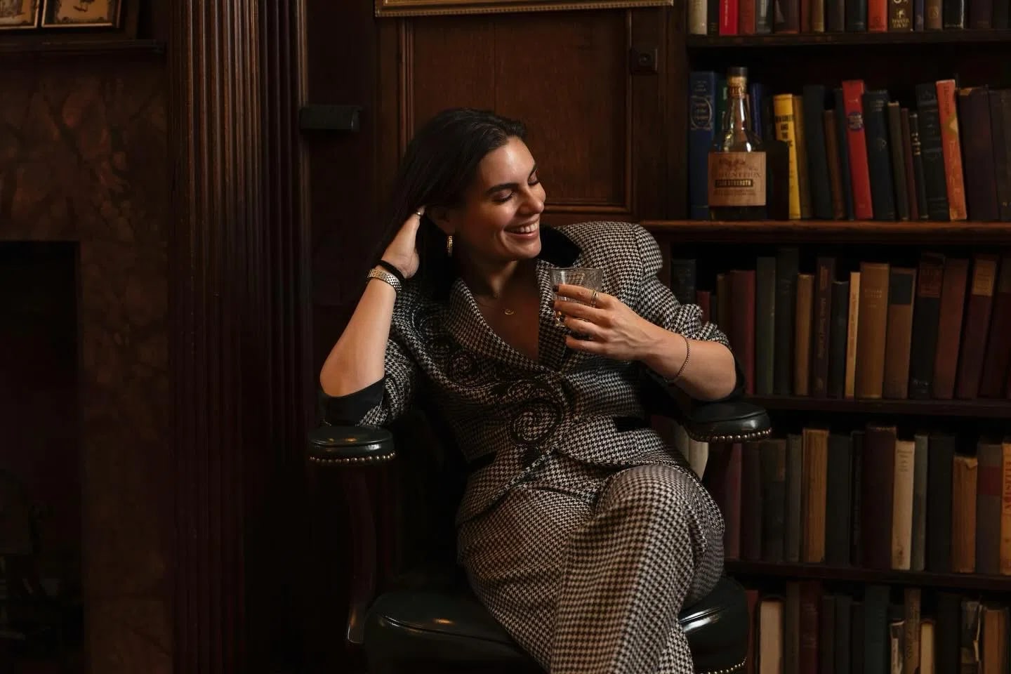 A smiling woman with dark hair sits in a vintage leather armchair in a cozy, dimly lit library, holding a short glass with a beverage. She is wearing a stylish black and white houndstoop skirt suit or pantsuit featuring a dark velvet collar and intricate black swirl detailing on the front. She rests her right hand against her head while looking downward and laughing. In the background, floor-to-ceiling wooden bookshelves are tightly packed with multi-colored antique books, and a bottle of liquor sits on an upper shelf, enhancing the classic, academic atmosphere.