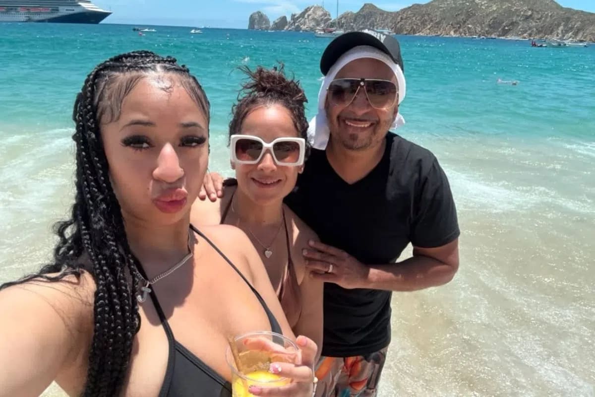A sunny, eye-level selfie captures three people smiling together on a tropical beach with turquoise water and rocky cliffs in the background. In the foreground, a young woman with long black braids and a black bikini top holds a clear plastic cup filled with a yellow drink, leaning into the frame. Standing close behind her is a woman with her hair up, wearing large white-rimmed sunglasses and a neutral-toned swimsuit, who smiles warmly toward the camera. To her right, a man in a black T-shirt, aviator sunglasses, and a black baseball cap worn over a white towel or fabric smiles broadly, completing the group. The ocean water fades from a vibrant turquoise to a frothy white shore around their ankles, while a large cruise ship and small boats are visible in the distance under a clear blue sky.