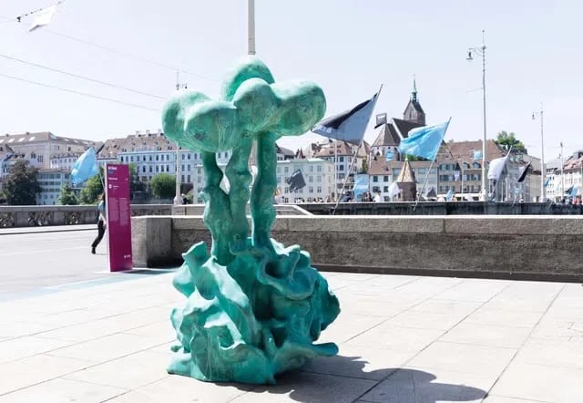 A turquoise abstract tree-like sculpture from Parcours 2026 installed on a paved plaza with historic Basel buildings  Credit: Photo: Art Basel / Parcours 2026  Place: In "The Street Pushes Back" section, after "with one installation on view across the river near Bankverein." Visitors to Art Basel 2026 will see a public artwork by Albanian Prime Minister and artist Edi Rama. His bronze sculpture, Untitled (2026), is on display in the “Parcours” public art section at the end of Clarastrasse, overlooking the Rhine River. 