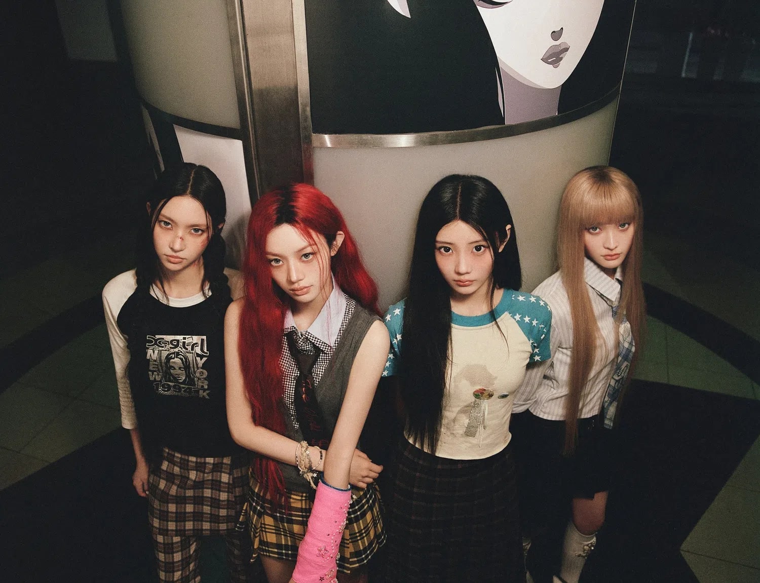 A wide, overhead shot captures the four members of the K-pop group ILLIT standing closely together, leaning against a curved architectural structure adorned with an illustration. They all look directly upward into the camera with serious, intense expressions. The member on the far left wears a black graphic tee with a "X-girl" print paired with dark plaid pants. The second member has vibrant red hair and a pink cast on her left arm, dressed in a grey sweater vest over a white collared shirt and a yellow plaid pleated skirt. The third member sports a cream-colored raglan tee with blue star-patterned sleeves and a dark plaid skirt. The member on the far right has long blonde hair with blunt bangs, wearing a striped button-up shirt, a tie, and a dark skirt. The overall aesthetic is moody and nostalgic, cast in warm, muted lighting.