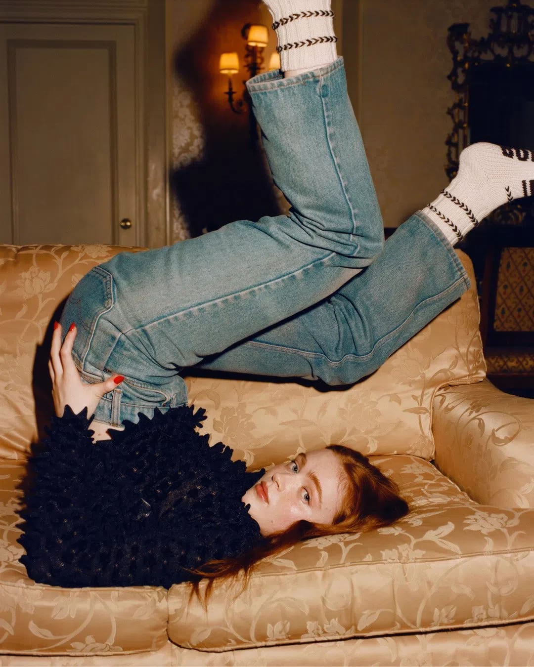 A young woman with long, vibrant red hair poses playfully upside down on a plush, gold damask-patterned sofa, her legs elevated high in the air while wearing mid-wash blue denim jeans paired with white, patterned crew socks. She wears an intricate, heavily textured navy or black long-sleeved top covered in small, 3D fabric embellishments or petal-like structures that give it a unique tactile depth. Resting on her back with her head tilted toward the camera, she gazes directly forward with a soft, neutral expression, accented by a bright red manicure on her hand resting near her hip. The indoor setting features a vintage aesthetic with warm, subdued lighting, highlighted by a glowing wall sconce in the background and a classic wooden paneled door, casting dramatic shadows that enhance the cozy, editorial feel of the photograph.