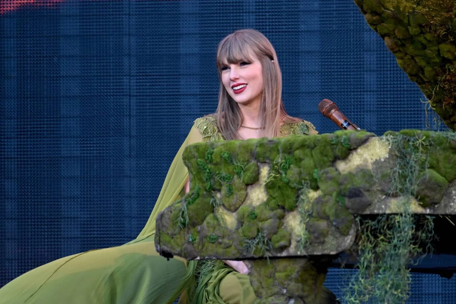 Taylor Swift sits smiling at a moss-covered grand piano on an outdoor stage during a concert performance. She is wearing a flowing, olive green gown with a matching cape and delicate lace detailing on the shoulders. Her blonde hair is styled straight with bangs, and she wears her signature bright red lipstick. The piano is heavily decorated with realistic green moss and hanging faux foliage, blending into a large mossy structure on the right. A wooden microphone stands near the piano keys. The background consists of a large, dark textured LED video screen showing a faint pattern.