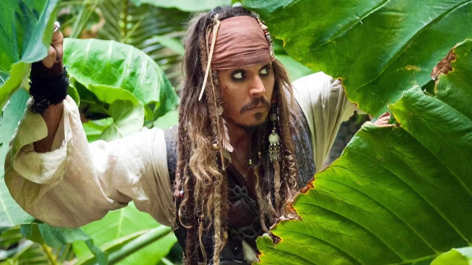 An eye-level shot captures a man dressed as a pirate peeking cautiously through large, vibrant green tropical leaves. He has long, dark dreadlocks adorned with beads and trinkets, a faded red bandana wrapped around his head, and dark eyeliner defining his intense expression. He wears a loose, cream-colored, long-sleeved shirt with wide sleeves under a dark textured vest. His right arm is raised, hand covered by a dark, fingerless glove as he pushes aside dense foliage to look toward the right side of the frame, appearing alert and stealthy within a lush jungle environment.