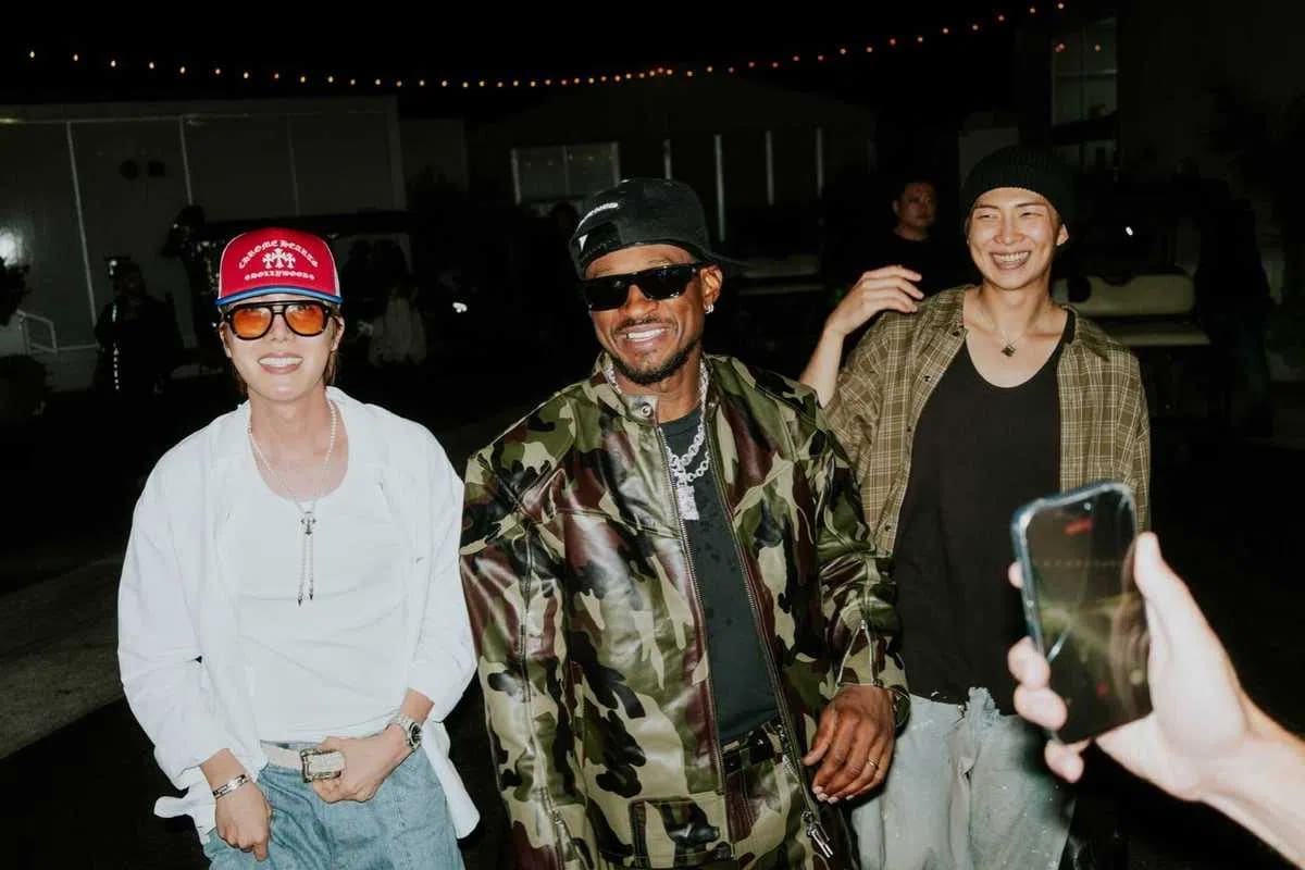 An image captured at night shows BTS members J-Hope and RM smiling and laughing alongside American singer Chris Brown. On the left, J-Hope wears a vibrant red trucker hat, tinted sunglasses, a loose white button-up shirt, and baggy light-wash jeans while flashing a wide smile. In the center, Chris Brown sports a camo-print leather jacket over a black top, a black baseball cap, dark sunglasses, and chunky silver chains. To the right, RM beams in a relaxed, casual outfit consisting of an open brown plaid flannel shirt over a black t-shirt and a dark beanie. The background shows an outdoor venue setting illuminated by string lights, with a hand holding up a smartphone in the foreground to snap a photo of the trio. 
