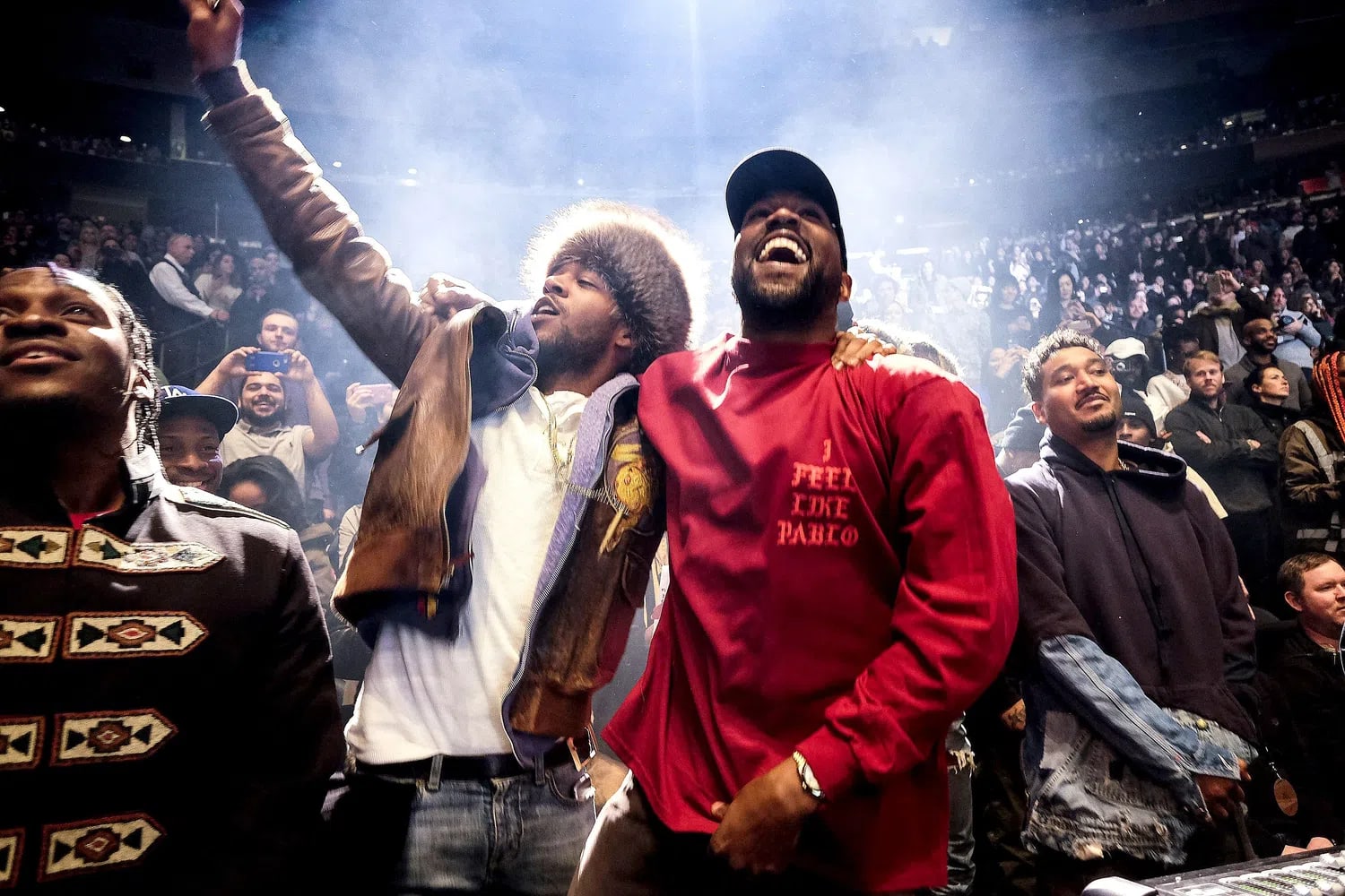 An image captures Kanye West and Kid Cudi sharing a moment of triumph during the listening party for West’s album The Life of Pablo at Madison Square Garden. Standing side-by-side in the midst of a packed, brightly illuminated stadium crowd, West laughs broadly while wearing a red long-sleeved shirt with the text "I FEEL LIKE PABLO" printed on the chest and a black cap. Beside him, Kid Cudi extends an arm high into the air while singing or shouting enthusiastically, dressed in a white t-shirt under a heavily layered brown jacket with a prominent fur-trimmed hood. To their left, rapper Pusha T is visible in profile looking upward, and behind them, an expansive audience fills the arena seats, captured under dramatic spotlighting that creates a hazy, energetic atmosphere.