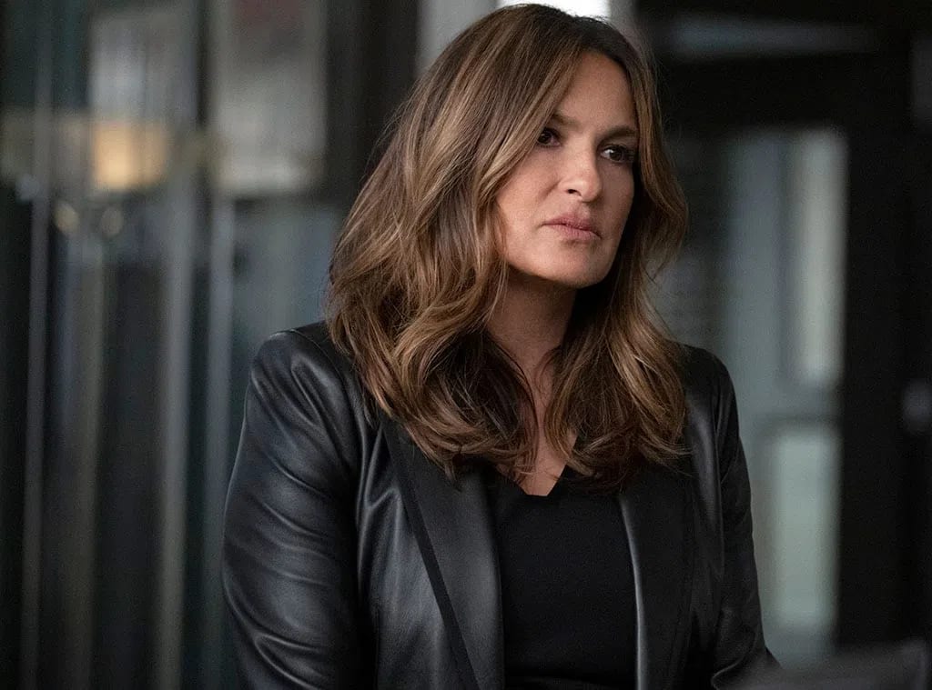 An image showcases American actress Mariska Hargitay, widely recognized for her iconic role as Captain Olivia Benson on the long-running NBC drama series Law & Order: Special Victims Unit, posing in a modern indoor setting with a serious, contemplative expression. Her mid-length, warm brunette hair is styled in voluminous, soft waves with subtle caramel highlights that frame her face elegantly. She wears a sophisticated ensemble featuring a sleek, tailored black leather blazer layered over a simple, matching black top, projecting a sharp, authoritative, and professional aesthetic. The background is softly blurred, hinting at an office or studio environment with vertical architectural elements and muted lighting that keeps the visual focus entirely on her compelling presence.