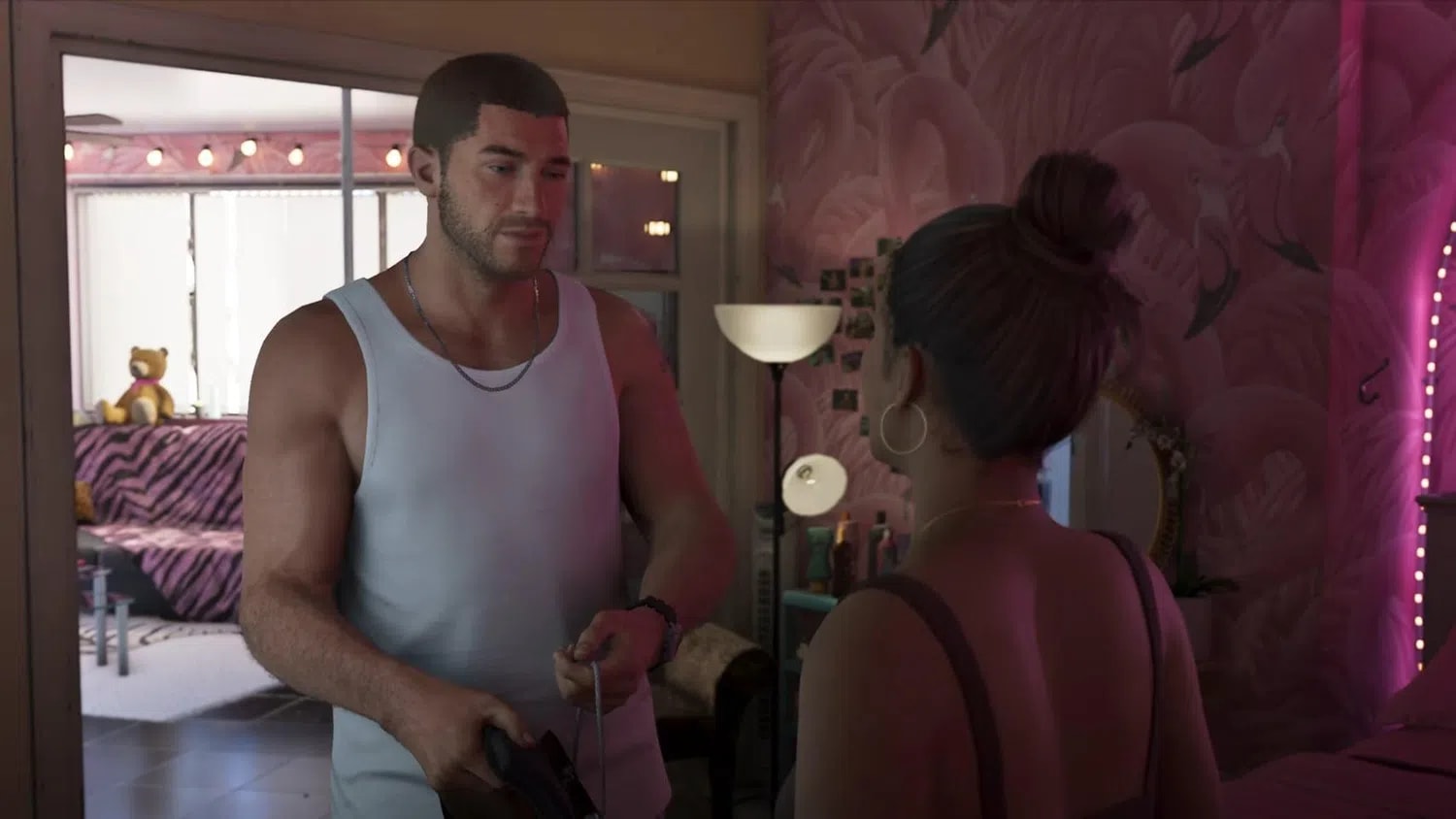 An intimate screenshot from the highly anticipated Grand Theft Auto VI video game trailer captures a tense yet romantic exchange between the two main protagonists inside a dimly lit room. On the left, Jason stands facing forward, wearing a white ribbed tank top and a simple silver chain necklace, looking down with a somber expression as he holds what appears to be a firearm. Lucia is positioned on the right with her back mostly to the camera, her hair tied up in a neat bun, sporting large hoop earrings and a dark strap top while staring intensely at him. The room's background features a vibrant pink wall adorned with flamingo patterns, strings of warm fairylights stretching across the ceiling, and a cozy couch with a zebra-print throw blanket in the adjacent room, creating a striking contrast between the domestic setting and the criminal undertones of their conversation.