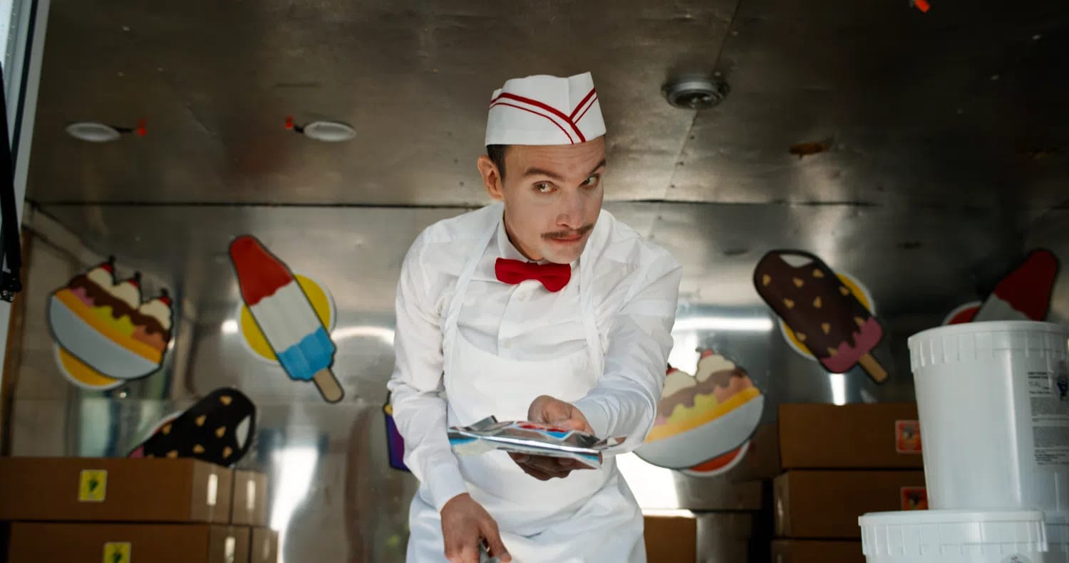 An official production promotional image from director Eli Roth's unrated horror-slasher film Ice Cream Man features actor Ari Millen portraying the menacing title character inside his dark, metallic ice cream truck. Leaning forward with an unsettling, wide-eyed stare and a sinister smirk, the slender vendor has a thin brown mustache and wears a vintage-style, crisp white uniform consisting of a long-sleeved button-up shirt, a matching white apron, a bright red bow tie, and a white paper soda jerk hat accented with red stripes. He extends his hand forward toward the camera, holding out a silver-wrapped frozen treat packet. In the softly blurred background, the interior of the food truck is visible, decorated with colorful, cartoonish graphics of frozen novelties including a rocket pop, an ice cream sundae bowl, and a chocolate-dipped ice cream bar with sprinkles, alongside stacked cardboard supply boxes sitting on the floor.