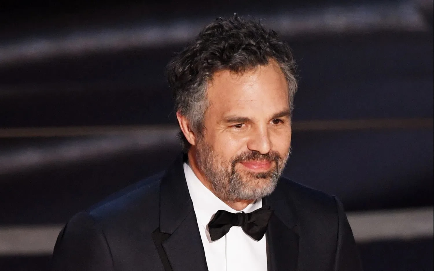 An older, middle-aged man with salt-and-pepper hair and a matching short beard smiles gently while posing for a photograph. He is wearing a formal black tuxedo jacket with sleek satin lapels over a crisp white button-down dress shirt. A classic black bowtie is neatly fastened at his collar. His dark curly hair is styled naturally with some volume, and his warm brown eyes look slightly off-camera. The background is a softly blurred, dark, textured surface with horizontal lines, suggesting an indoor red carpet or awards ceremony venue.