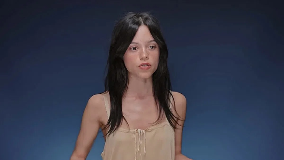 An over-the-shoulder medium shot features actress Jenna Ortega speaking directly to the camera against a solid navy blue studio backdrop. She has middle-parted, dark shoulder-length hair with soft curtain bangs framing her face. She wears a minimalist, beige sleeveless camisole top with delicate tie-front detailing and lace trim around the neckline. Her expression is focused and animated mid-speech, lit by soft, even studio lighting that highlights her features. An interview still shows actress Jenna Ortega from the waist up against a solid, dark blue gradient backdrop. She has dark, shoulder-length hair with soft curtain bangs framed around her face, and she is looking slightly to her right with an open mouth as if speaking. She wears a lightweight, sleeveless beige or light tan top featuring delicate thin straps and a small tie detail at the center of the scoop neckline.