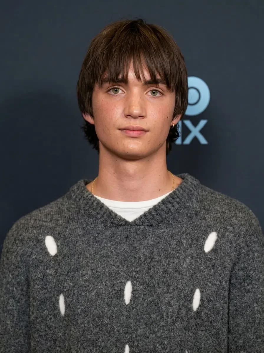 The image features actor Aran Murphy, son of Cillian Murphy, posing at a red carpet event against a dark blue background adorned with a partial "DX" logo. He is looking directly at the camera with a subtle, neutral expression, sporting a textured brown bowl-cut hairstyle with long bangs framing his eyes, and a hint of a small gold hoop earring in his left ear. He is wearing a cozy, thick grey wool knit sweater featuring an open, slightly distressed texture with visible white spots or under-layer accents peeking through, worn over a simple white crew-neck undershirt and a thin gold chain necklace.