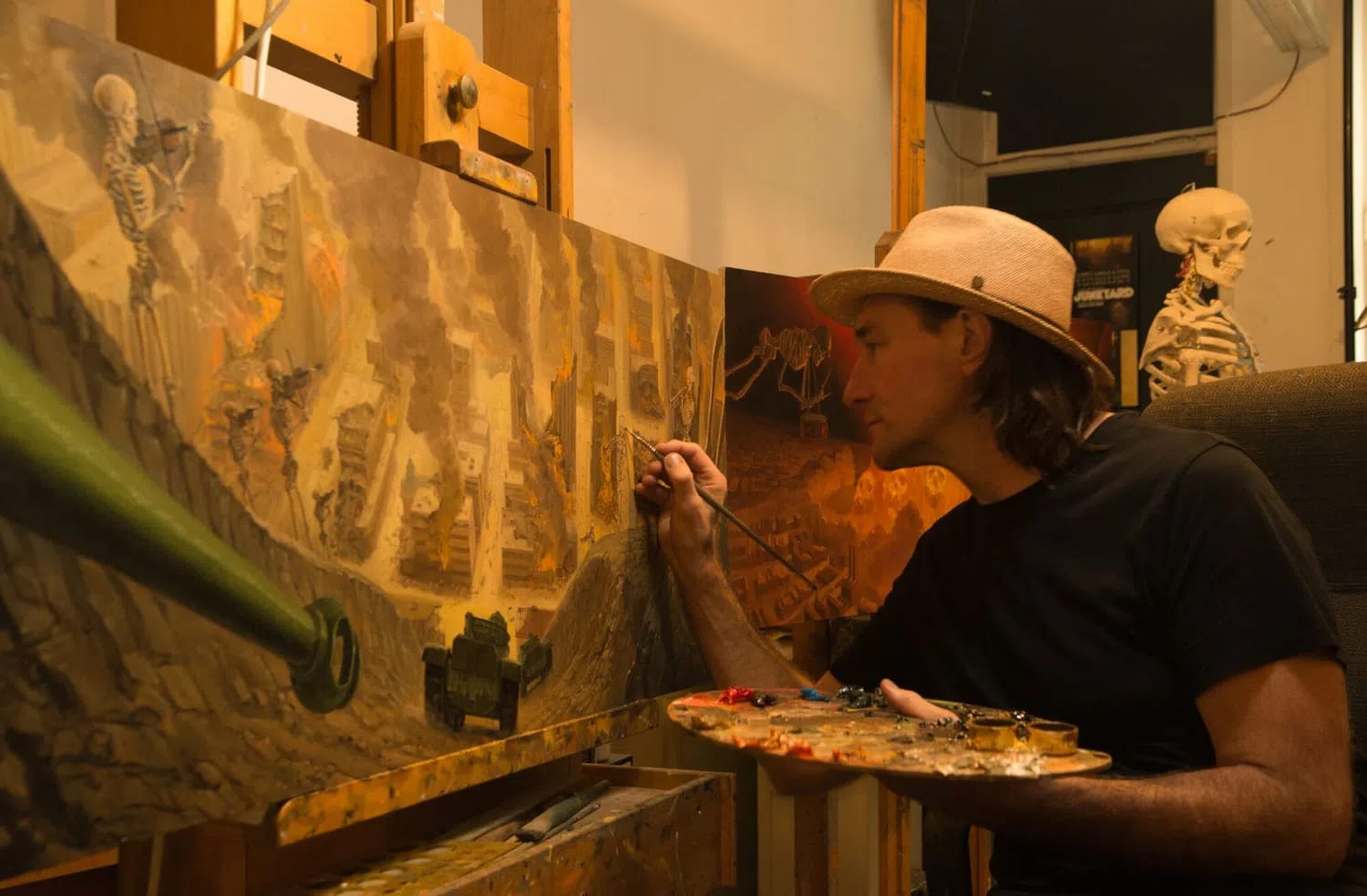 Artist Hisko Hulsing is captured in his studio, focused on a detailed oil painting of a war-torn cityscape filled with smoke, fire, and a skeletal figure playing a violin. Wearing a tan fedora and a black T-shirt, he sits in profile, holding a fine-tipped brush to the canvas with his right hand while balancing a wooden palette smeared with vibrant paints in his left. In the warm-toned background, another painting and a medical skeleton model are visible, adding to the atmosphere of a dedicated creative workspace.
