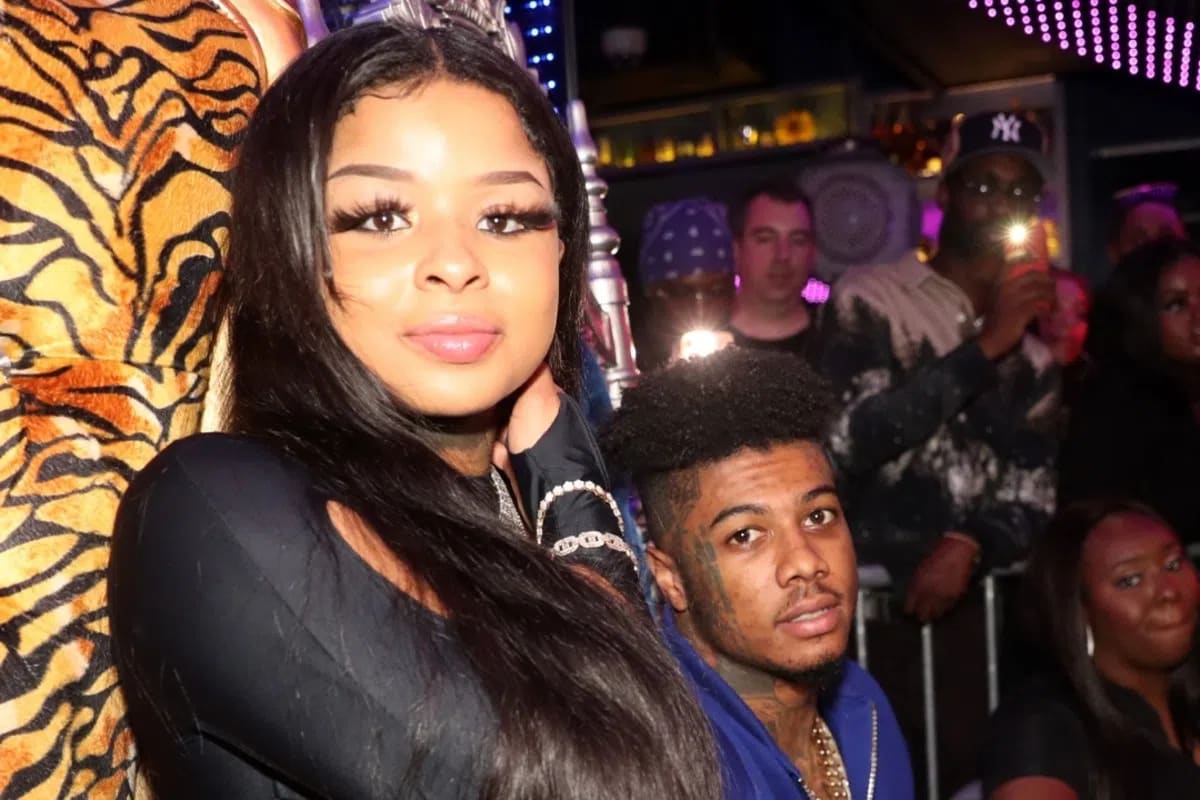 A medium close-up shot captures media personalities Chrisean Rock and Blueface inside a crowded, dimly lit nightclub venue. Chrisean Rock stands prominently on the left side of the frame, looking directly into the camera with long, dark straight hair draped over her shoulder and a subtle smile. She wears a long-sleeved black top and a silver metallic chain bracelet on her wrist. Behind her left shoulder, Blueface stands slightly lowered in the frame, wearing a blue jacket with his characteristic facial tattoos visible. The background is filled with a dense crowd of clubgoers behind a metal security barrier, multiple camera flashes illuminating the dark space, neon purple lighting patterns on the ceiling, and a tiger-print patterned garment visible on the far left.