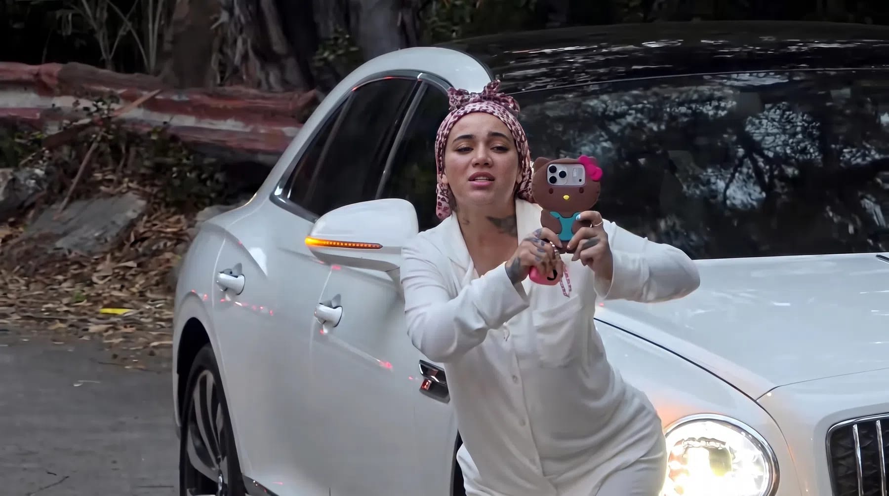 American media personality and rapper Danielle Bregoli, widely recognized as Bhad Bhabie, leans dynamically forward against a luxurious white Bentley Flying Spur while aggressively aiming her smartphone outward during a highly publicized confrontation in Hollywood. She is dressed in an off-white, long-sleeved loungewear set paired with a pink-and-white leopard-print headscarf wrapped tightly over her hair. Her expression is visually tense, marked by furrowed brows and parted lips as she fiercely records or gestures toward an off-screen group of live streamers. Her smartphone is prominently enclosed in a bulky, distinctive brown-and-pink Hello Kitty protective phone case. The luxury car behind her sits angled on a paved asphalt roadway, showing illuminated circular headlights and a glowing orange side-marker signal mirroring the chaotic, high-visibility energy of the scene. In the dimly lit background, natural foliage, overhanging branches, and a concrete roadside barrier provide a stark, unrefined contrast to the sleek design of the premium sedan.