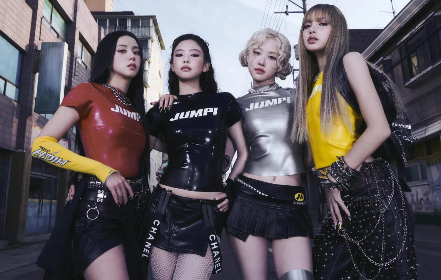 BLACKPINK stands firmly side-by-side in a gritty, industrial street setting under an open sky, radiating a powerful and modern aesthetic. The members pose in highly coordinated, edgy streetwear outfits featuring sleek, tight-fitting tops emblazoned with bold athletic branding. Jisoo commands attention on the left in a vibrant red latex shirt paired with a distinctive yellow arm sleeve. Next to her, Jennie wears a gloss-black "JUMPI" t-shirt over a structured, branded belt skirt. Rosé shines with pale blonde curls in a metallic silver mock-neck top and a micro pleated skirt, while Lisa anchors the right side of the frame in a bright yellow sleeveless top, sporting her signature long bangs and intricate heavy-metal chain detailing across dark pants.