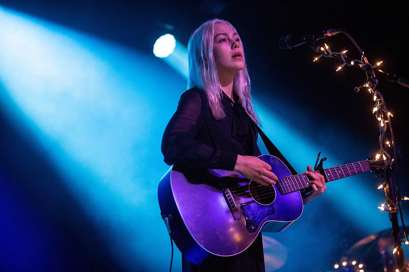 An eye-level shot captures singer-songwriter Phoebe Bridgers performing live on stage under intense blue and white spotlights. She has long, platinum blonde hair and wears a sheer black long-sleeved blouse with a high tie neck. She looks upward with an emotional expression while strumming a dark acoustic guitar with a prominent pickguard and a capo clamped on the neck. To her right, a black microphone stand is heavily wrapped in glowing, warm-toned fairy lights, creating a stark, moody contrast against the dark, hazy background of the venue.  Who is Phoebe Bridgers? Phoebe Bridgers (born August 17, 1994) is an American singer, songwriter, and musician known for her melancholic indie-folk and rock sound, confessional lyrics, and signature skeleton-suit stage attire. She has earned widespread critical acclaim and multiple Grammy nominations as a solo artist and as part of the indie supergroup Boygenius.