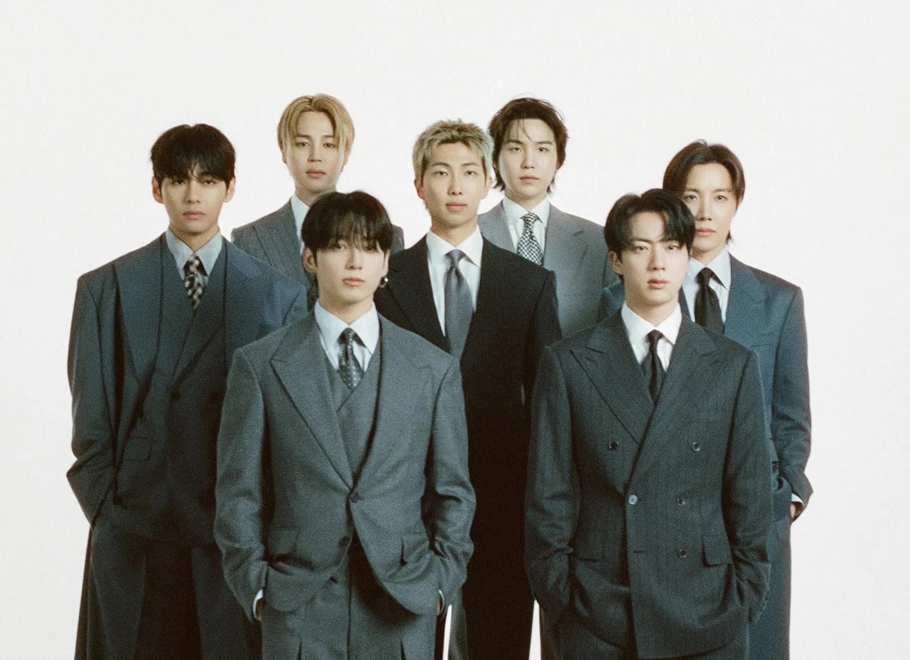 A group of seven young men, the globally acclaimed South Korean boy band BTS, pose together against a minimalist white studio background for a formal promotional poster. The members—Jungkook, Suga, J-Hope, RM, Jimin, V, and Jin—are arranged on stacked white rectangular platforms, dressed in sophisticated, monochromatic suits ranging from charcoal grey to deep black, paired with collared shirts and ties. Above them, a solid black banner prominently displays the text "ARTIST OF THE YEAR" in bold yellow lettering, while the bottom of the poster features a dark, cosmic outer-space graphic with the group's name "BTS" in large white font alongside official corporate logos for the MAMA Awards and Visa.