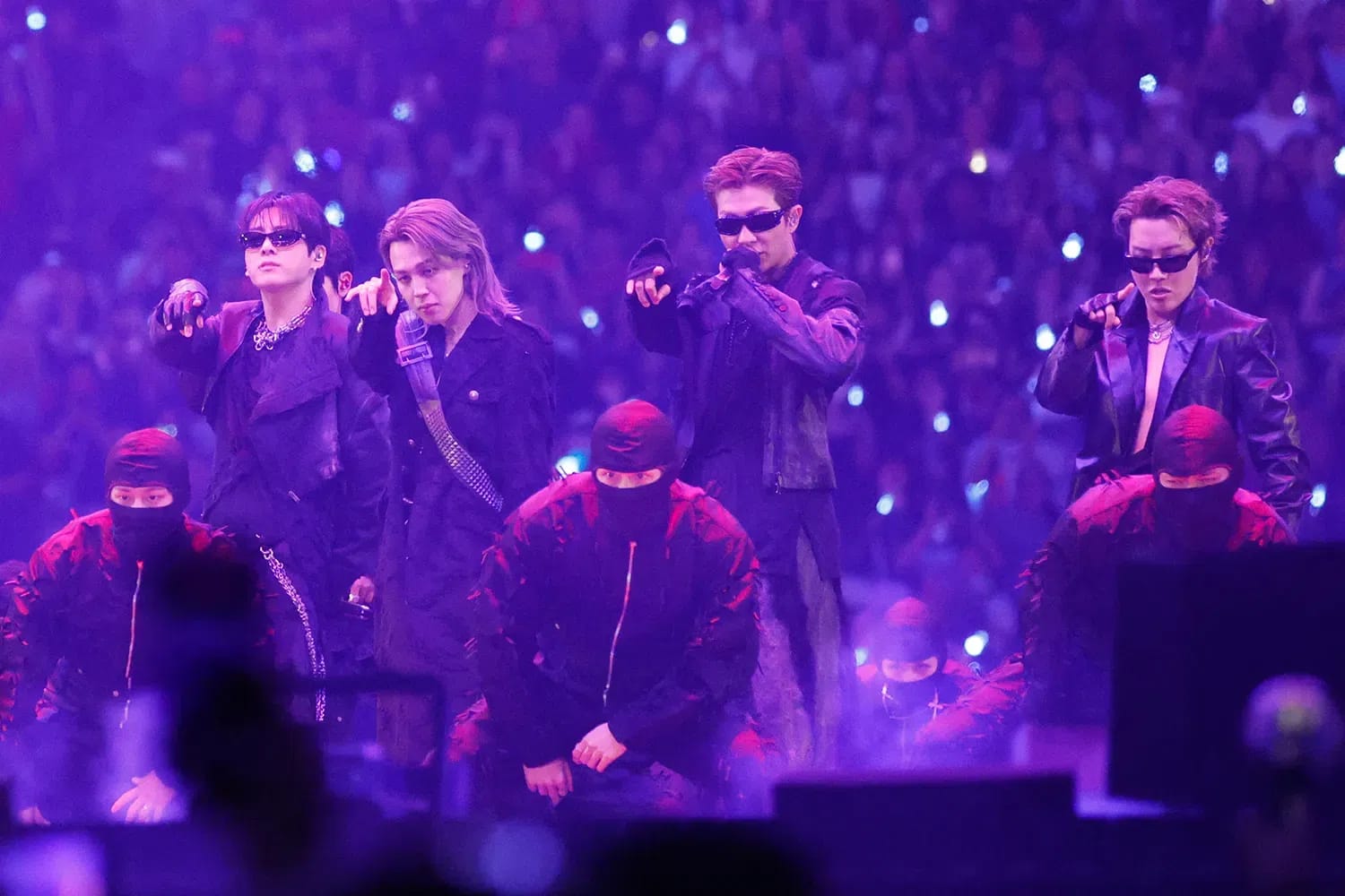 A wide-angle, concert-stage photograph captures four members of the South Korean boy band BTS performing under intense, vibrant purple lighting during a live stadium show. The performers stand side-by-side in a dynamic lineup, wearing stylized, dark edgy clothing, with three of them donning black sunglasses. From left to right, Jungkook gestures forward with his hand, Jimin looks intently toward the audience, RM holds a microphone up to speak while pointing forward, and J-Hope strikes a stylized pose on the right. In front of them, three backup dancers dressed in matching black balaclavas and dark jackets crouch low toward the stage floor. The background is filled with a vast, blurry crowd of fans holding glowing lightsticks, creating an immersive, high-energy concert atmosphere.